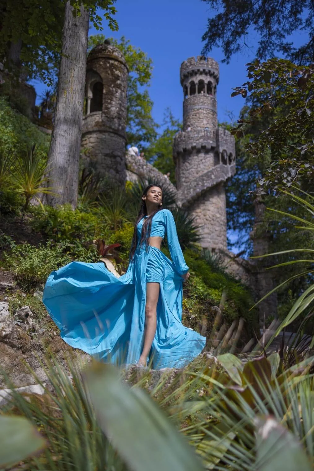 A woman in a flowing blue dress standing outdoors near a stone staircase with castle-like towers, surrounded by greenery and trees, under a blue sky.