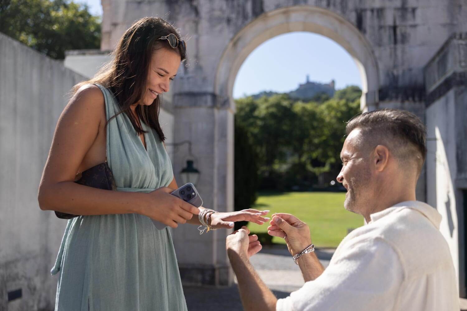 A woman is smiling as she receives a ring from a man outdoors near a stone archway, with green trees and a hilltop structure in the background on a sunny day.