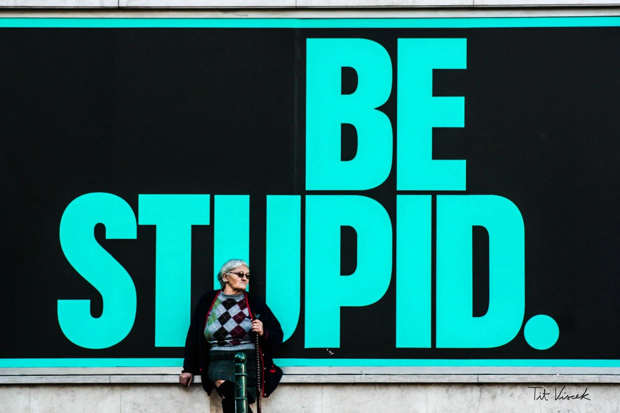 A large advertisement board that reads 'BE STUPID.' in bold turquoise letters on a black background, with an elderly woman wearing sunglasses, a sweater, and a skirt standing in front of it.