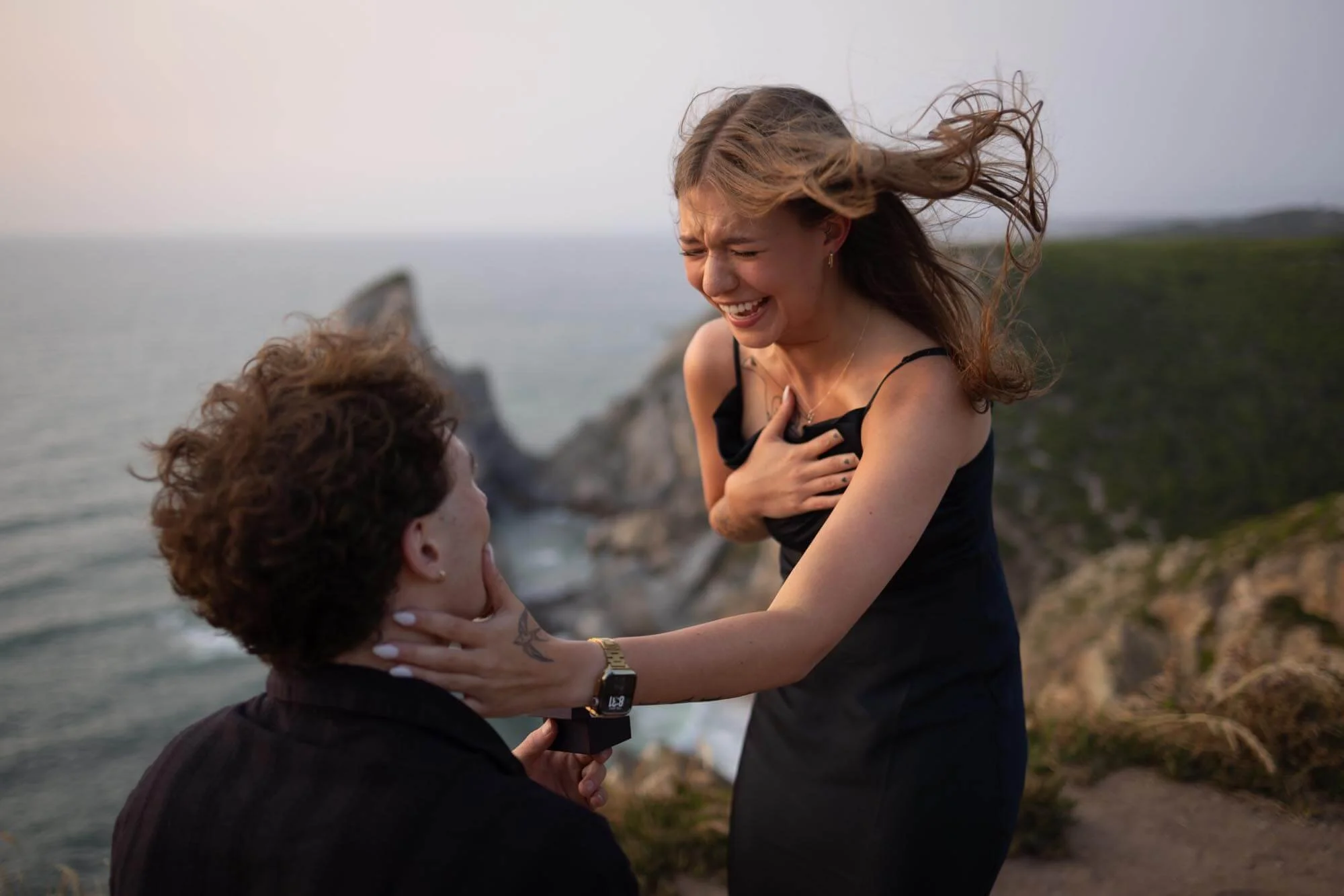 A woman in a black dress and a man with curly hair are near the coast, with the woman holding the man’s face and both smiling and laughing, with a rocky cliff and the ocean in the background.