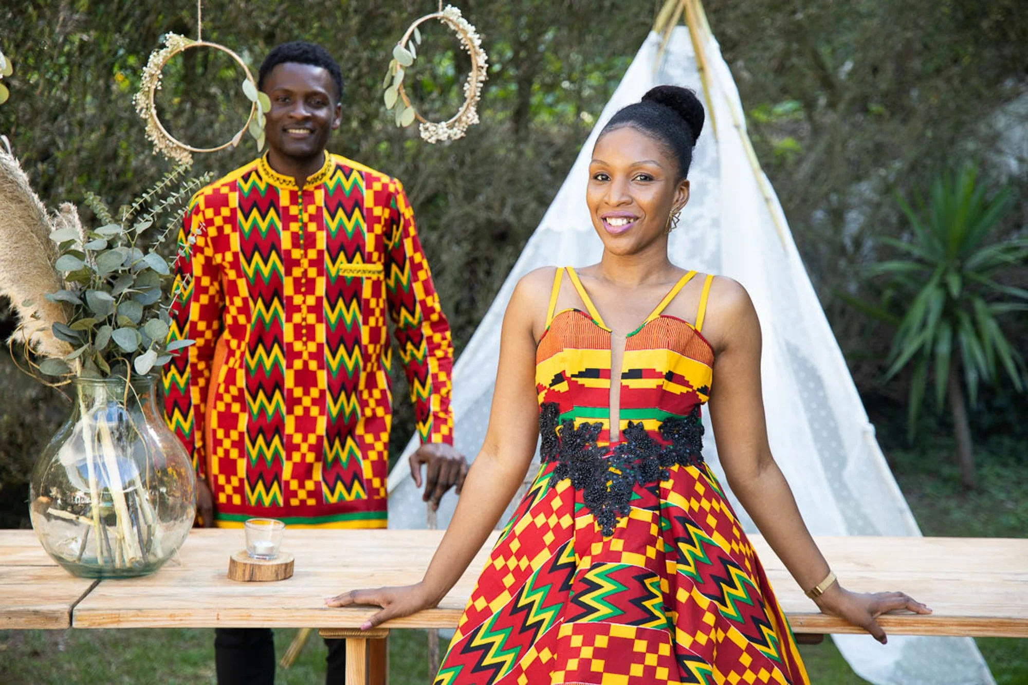 A smiling man and woman in colorful African traditional attire posing outdoors near a rustic wooden table with decorative plants and a teepee backdrop.