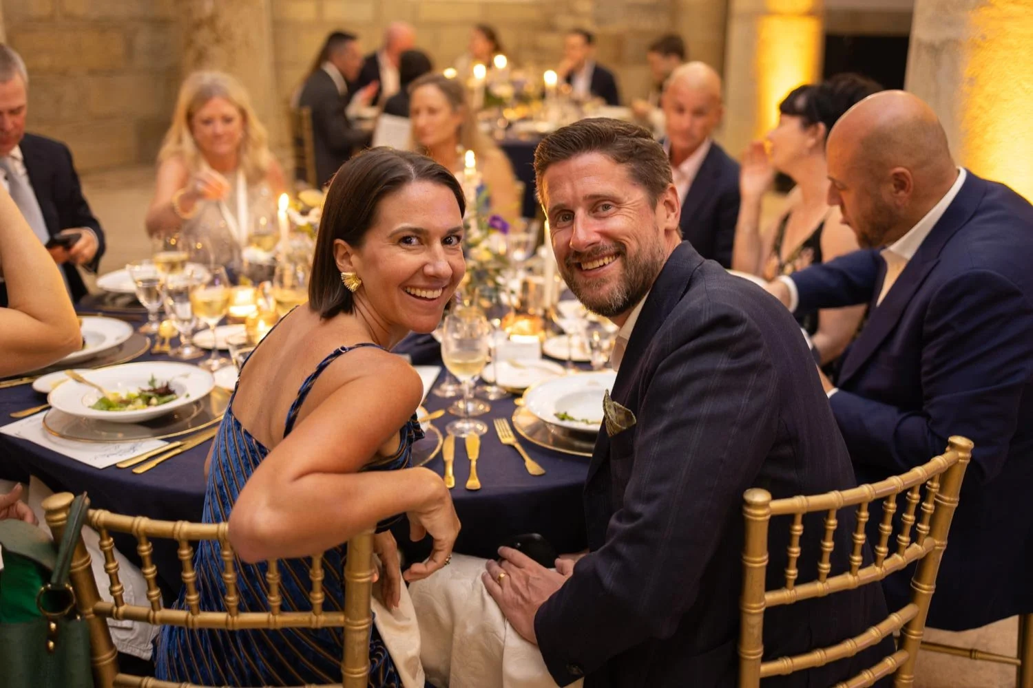 Smiling woman and man sitting at a formal corporate dinner table with elegant table settings, surrounded by other well-dressed guests in a decorated banquet hall. They are posing for an event photographer.