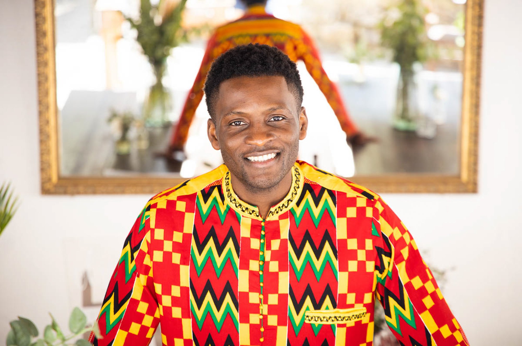 Smiling man in colorful traditional African clothing standing indoors with a mirror reflecting a person dressed in similar attire behind him.