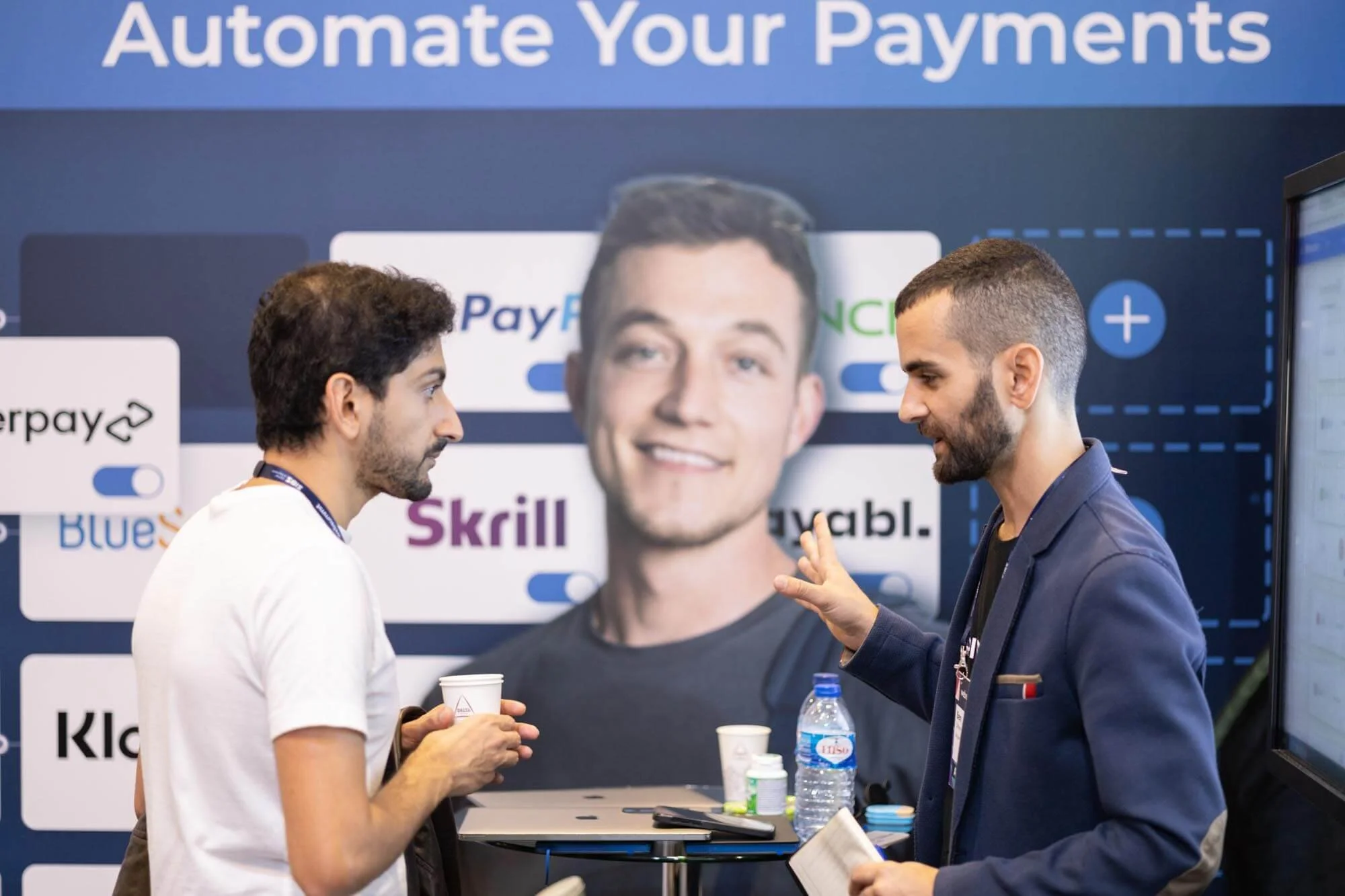 Two men engaged in a conversation at a conference booth promoting automatic payments, with a large display behind them showing logos of payment platforms like PayPal, Skrill, and Venmo, and the phrase 'Automate Your Payments'.