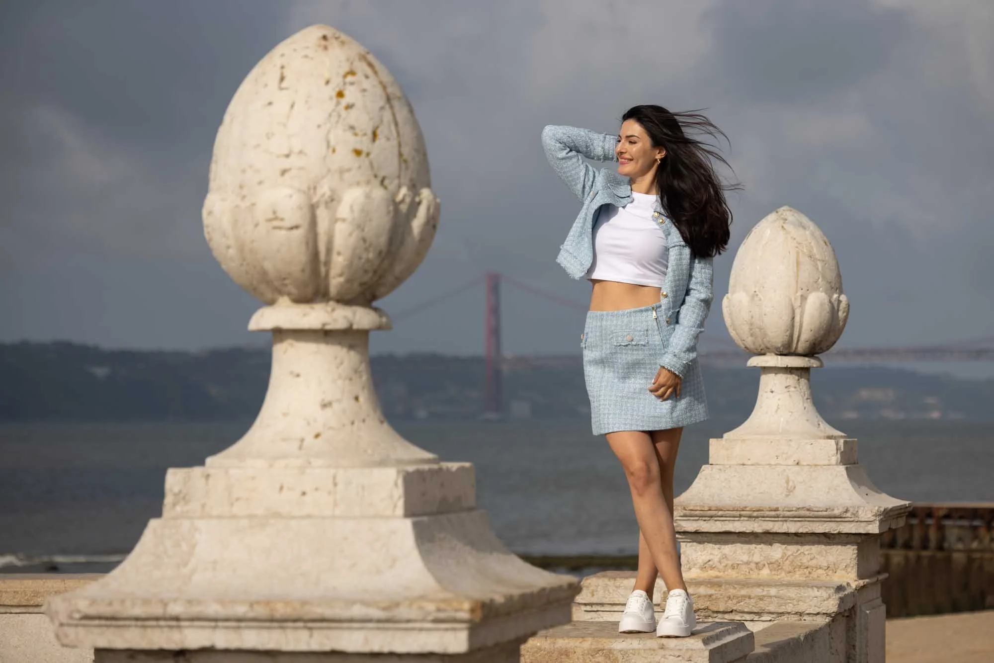 Beautiful woman is having a photoshoot at Lisbon river side. She has blue top and a skirt and crossed legs.