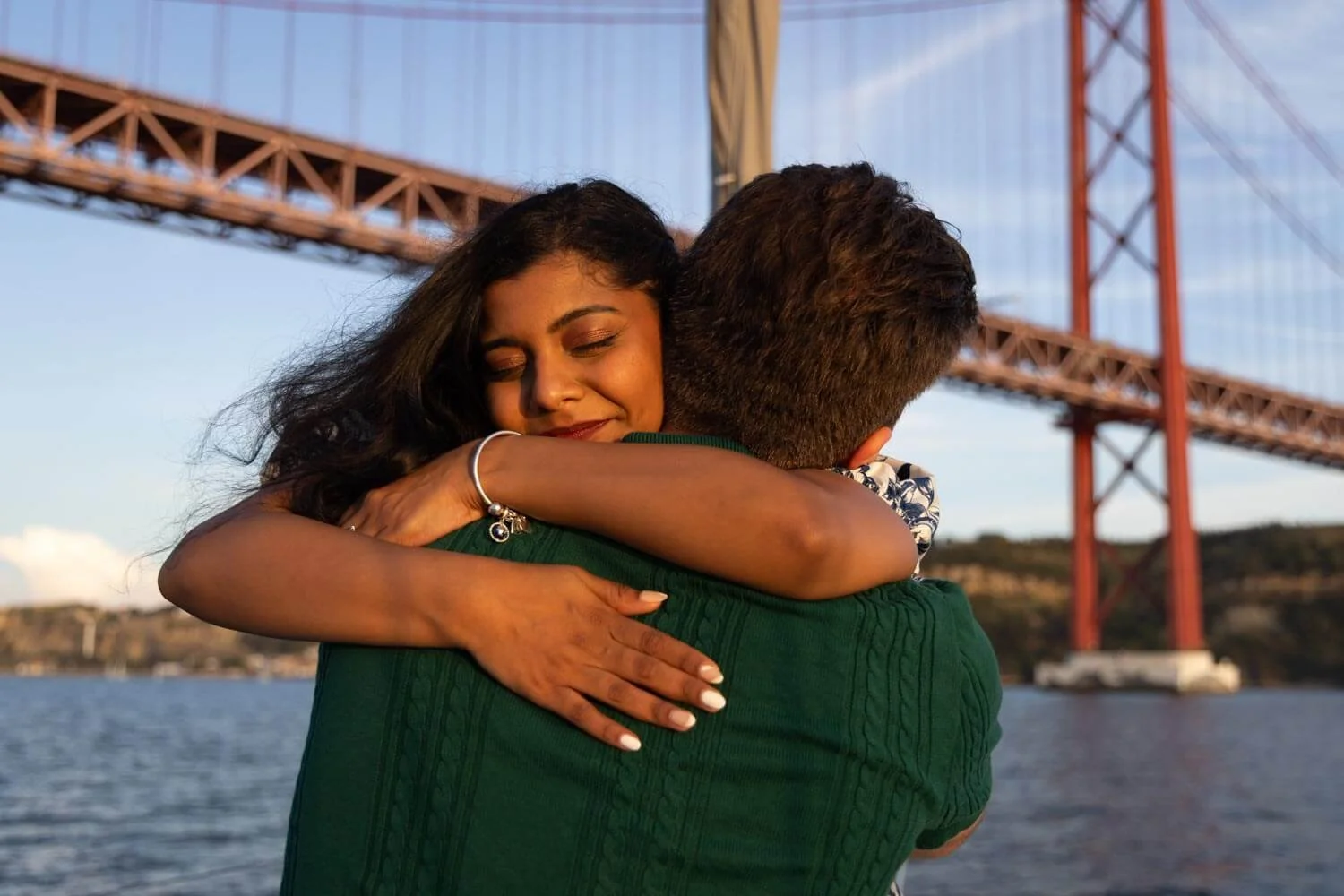 A woman with long dark hair and a bracelet hugging a man in front of a bridge over water.
