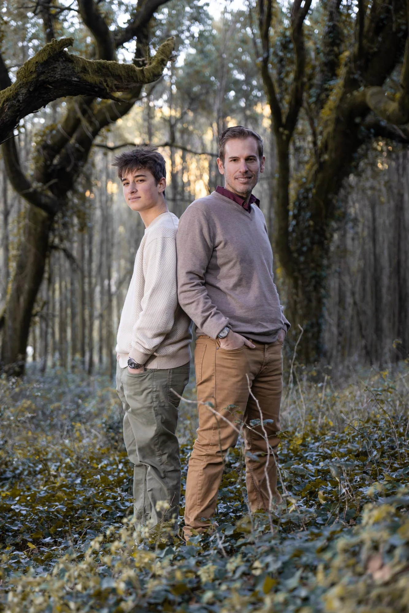 Two men standing back to back in a wooded forest at sunset, one young and one older, both wearing casual clothing.