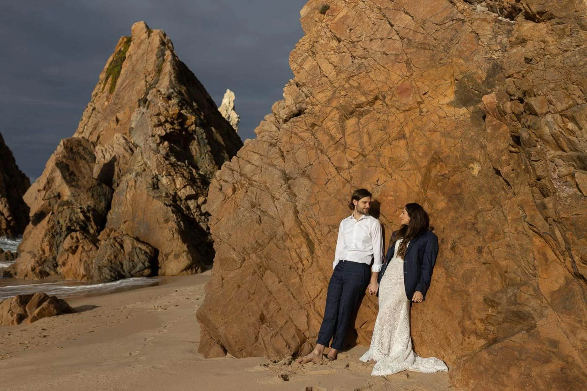 A man and woman standing on a sandy beach beside large rocky cliffs, with the man holding the woman's hand, both gazing at each other.