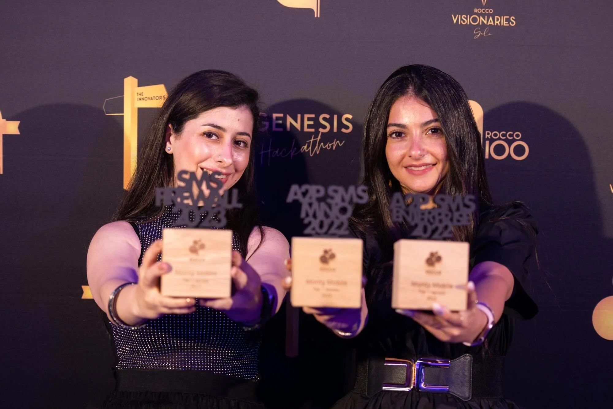 Two women holding awards at an event with a dark background featuring logos and text including "Genesis Hackathon" and "Rocco 100". They are smiling and facing the camera.