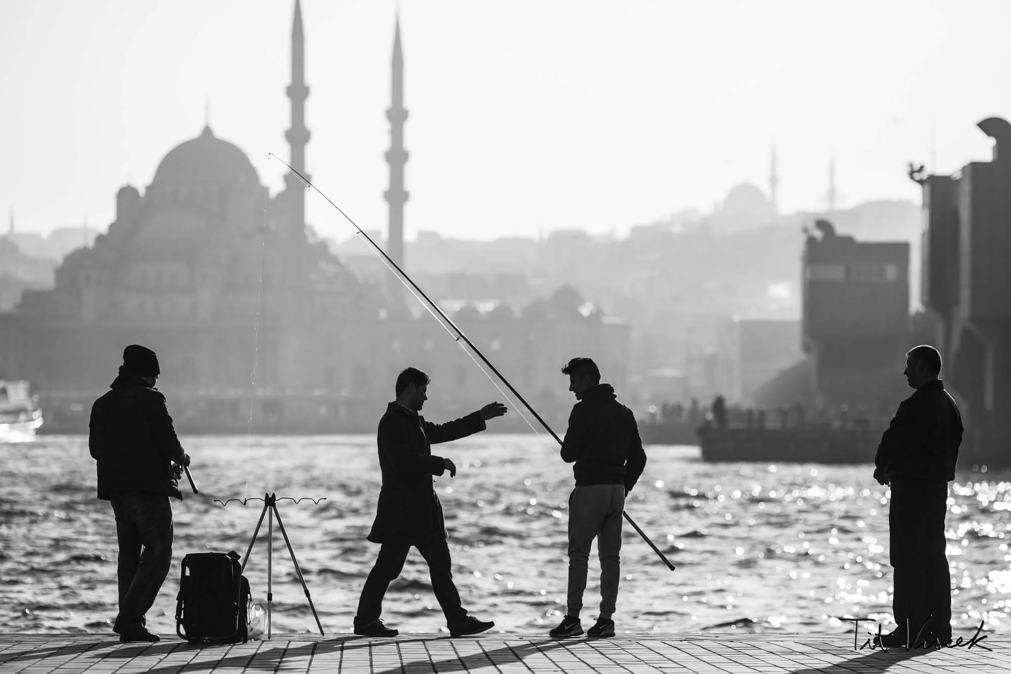 Four men standing by water, one fishing, with a mosque in the background, black-and-white photograph.