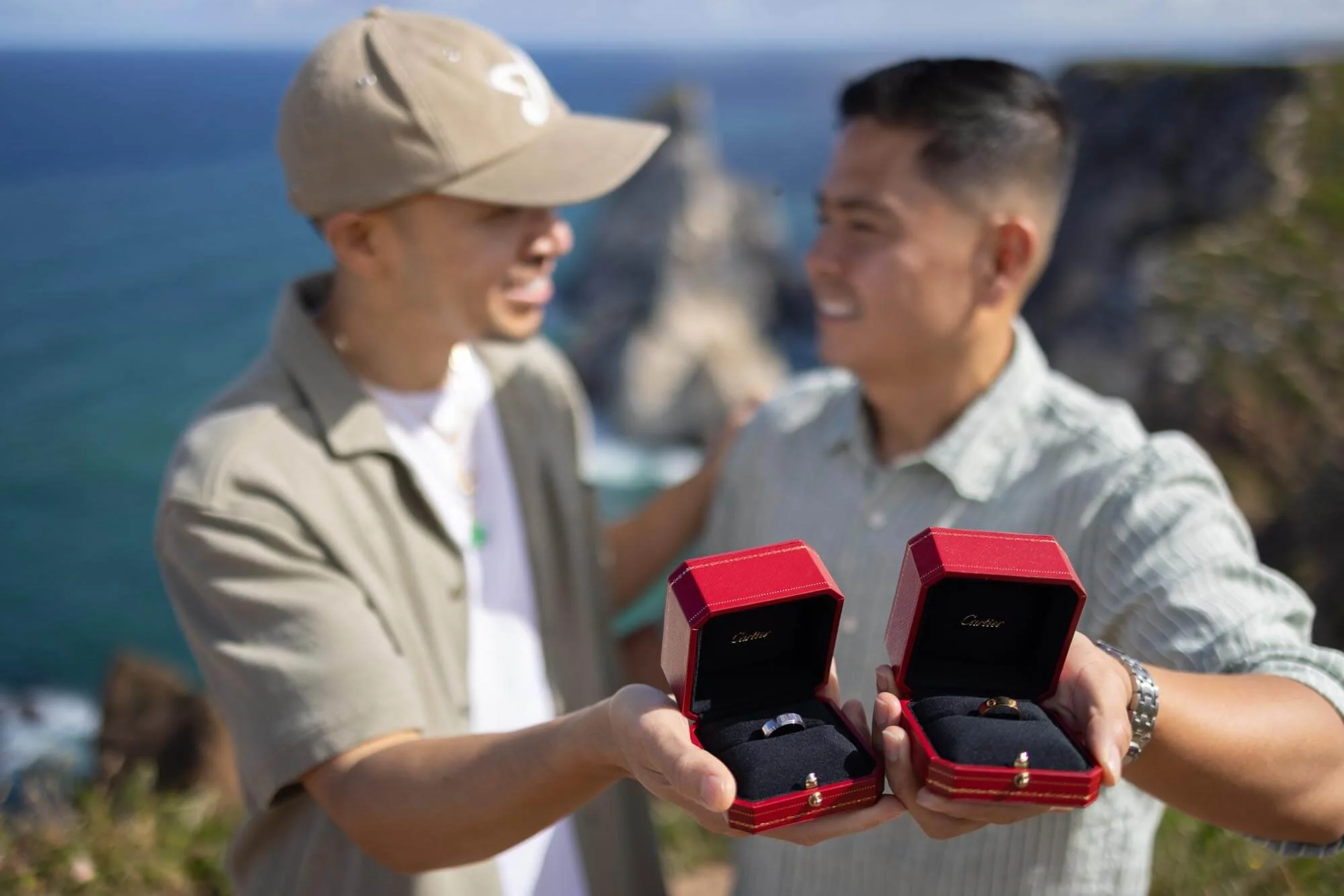 Two men exchanging jewelry in red rings boxes with a scenic ocean view in the background.