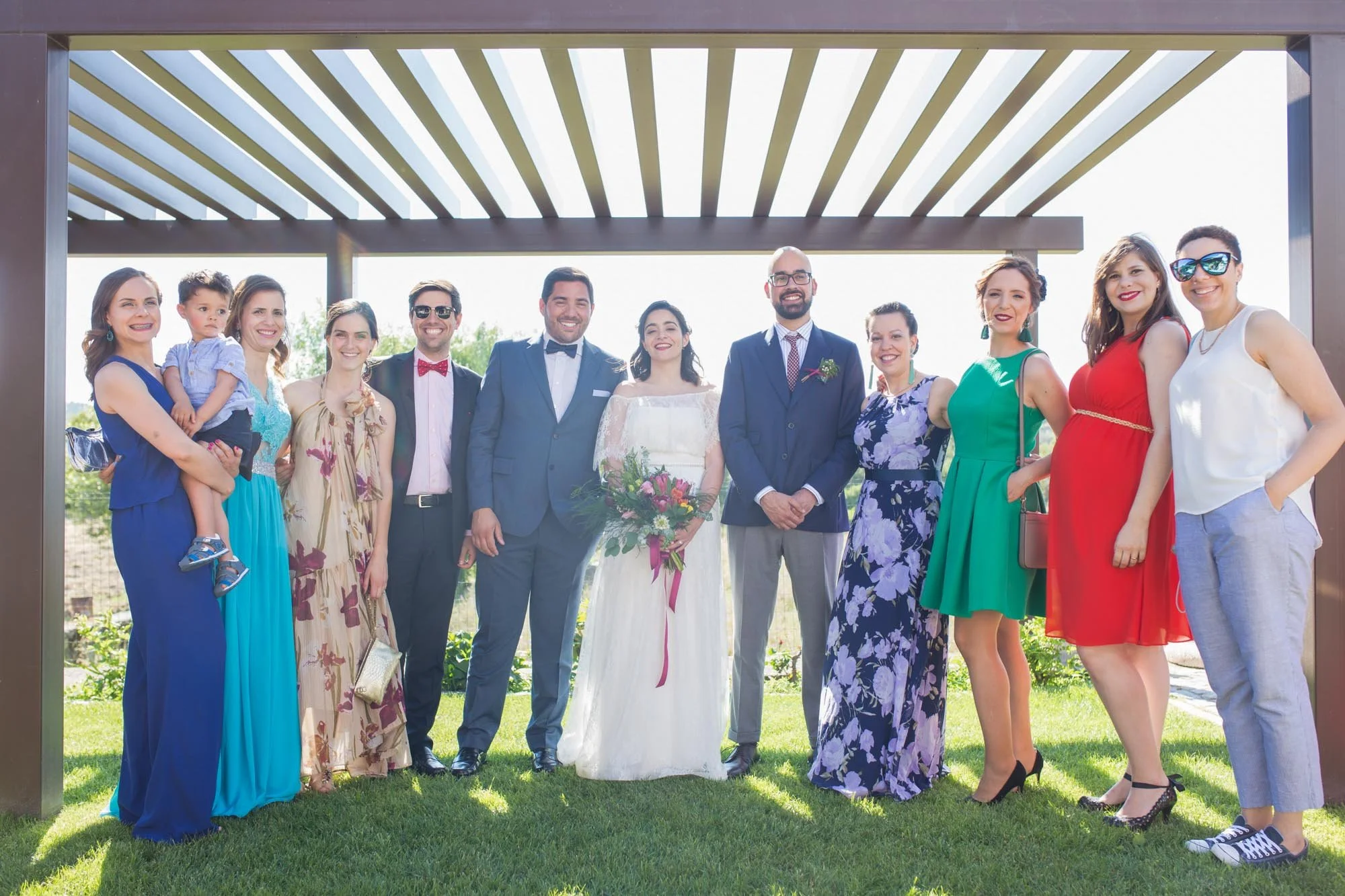 A group of people, including a bride in a white dress holding a bouquet, and wedding guests, standing outdoors under a pergola, smiling for a photo on a sunny day.