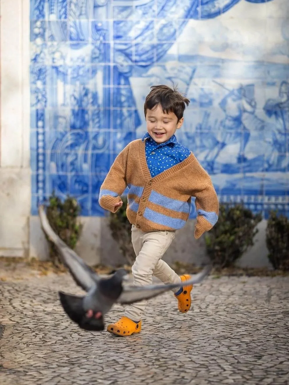 A young boy playing outdoors, running and smiling, wearing a brown and blue striped cardigan, beige pants, and orange Crocs, with a pigeon flying close to the camera, against a background of a blue mural.