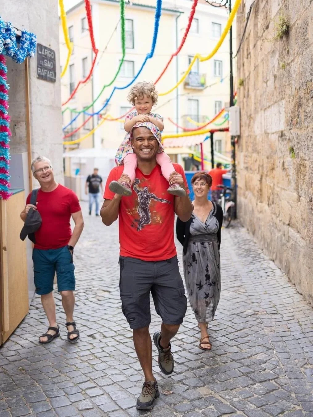 A man is carrying a young girl on his shoulders while walking down a cobblestone street, smiling. Two other adults are walking behind them, also smiling. The street is decorated with colorful streamers hanging overhead.