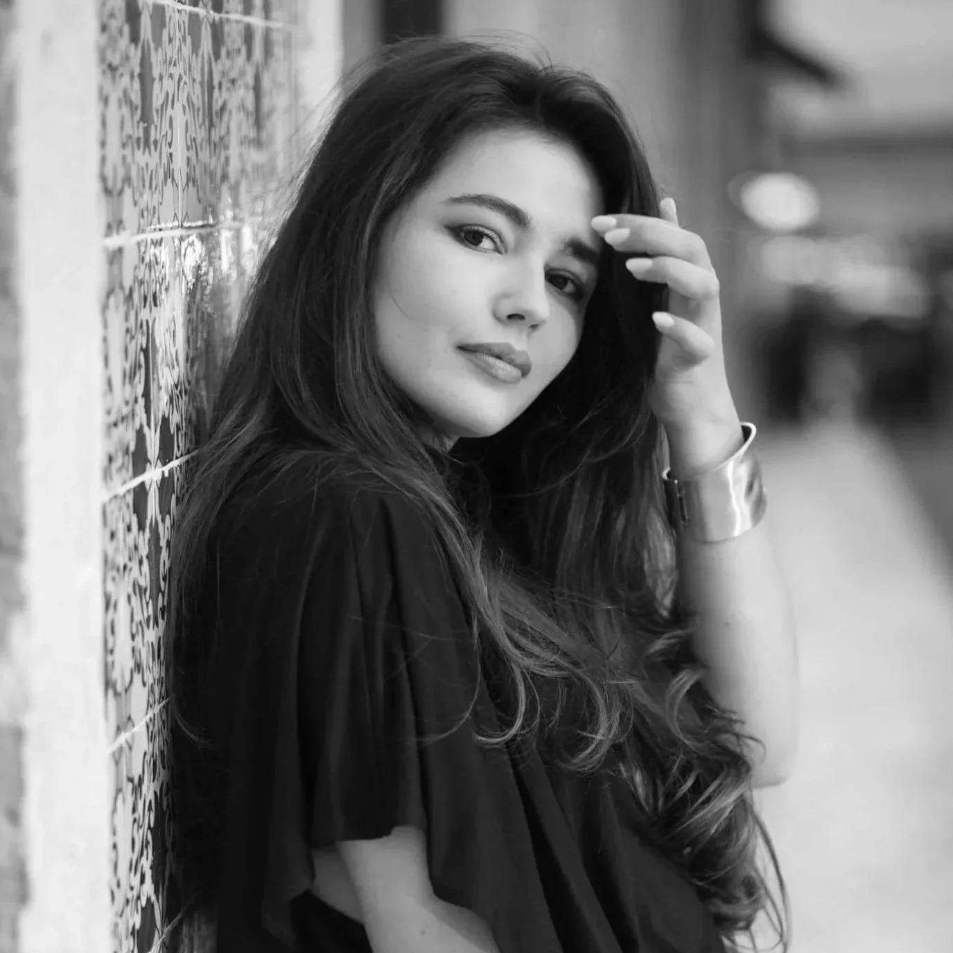 Black and white photo of a woman with long hair leaning against a tiled wall, she is looking at the camera and touching her hair.