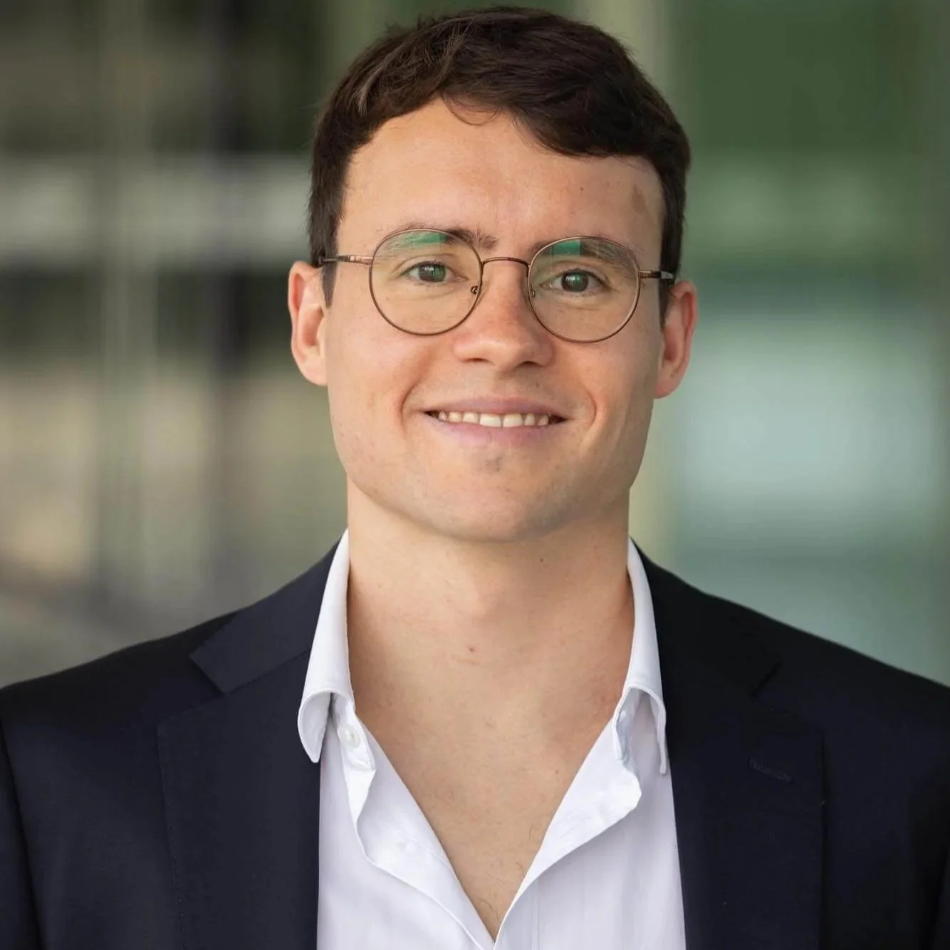 A young man with short brown hair, glasses, and light skin, wearing a white shirt and black blazer, smiling at the camera. The background is blurred.
