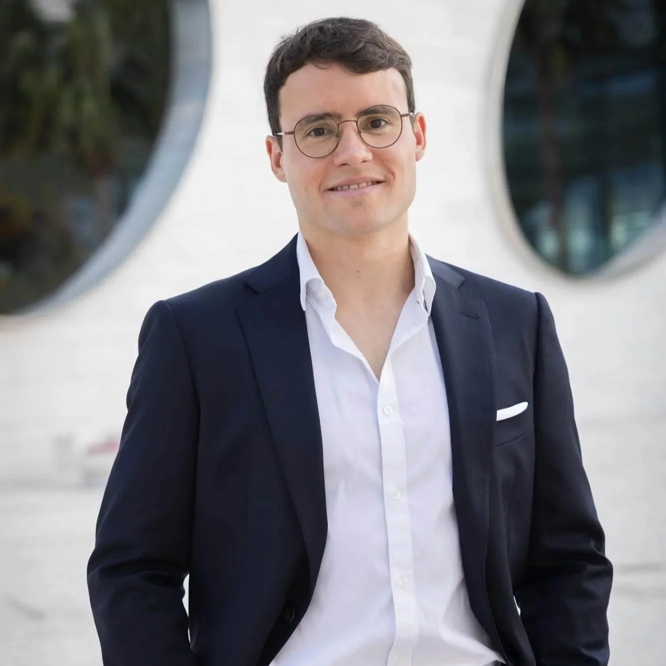 A man with short dark hair, wearing glasses, a white dress shirt, and a dark blazer, smiling outdoors with circular windows in the background.