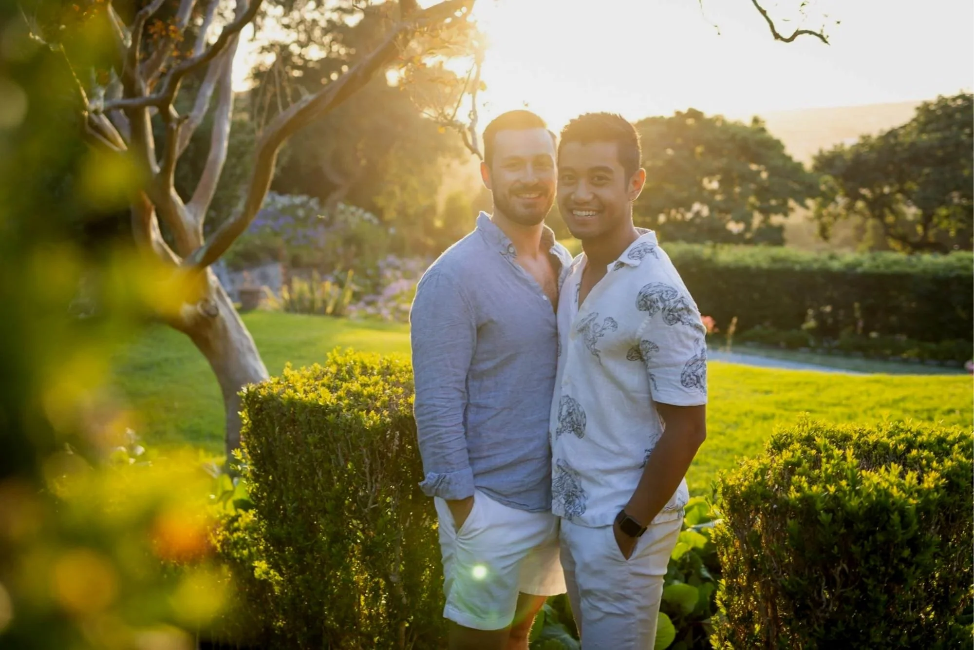 Two men standing close together outdoors during golden hour sunset, smiling, surrounded by greenery and trees.