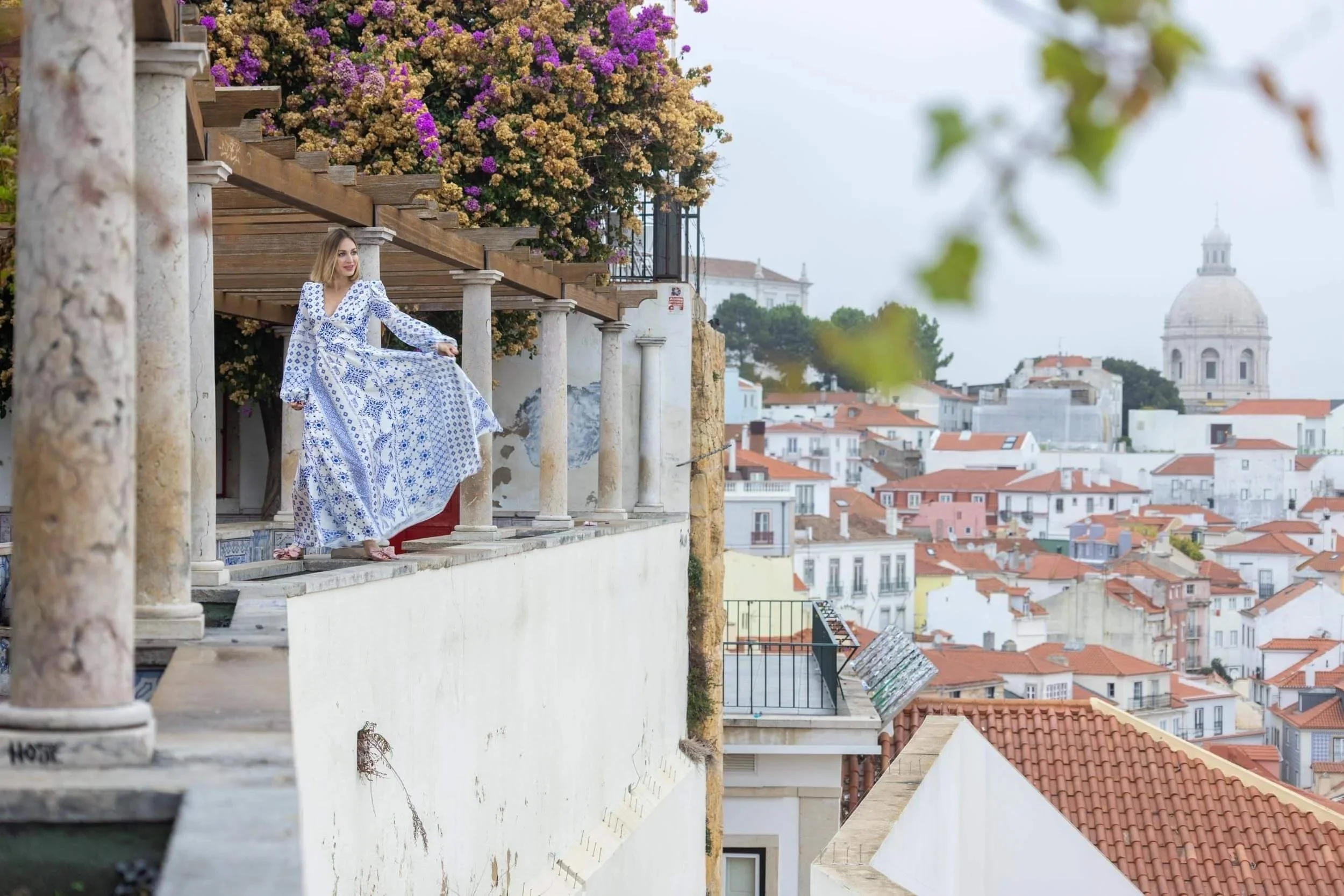 A woman in a blue and white patterned dress stands on a Lisbon viewpoint overlooking a cityscape with white buildings and red-tiled roofs, and a large domed building in the background, with some greenery and blooming flowers nearby.