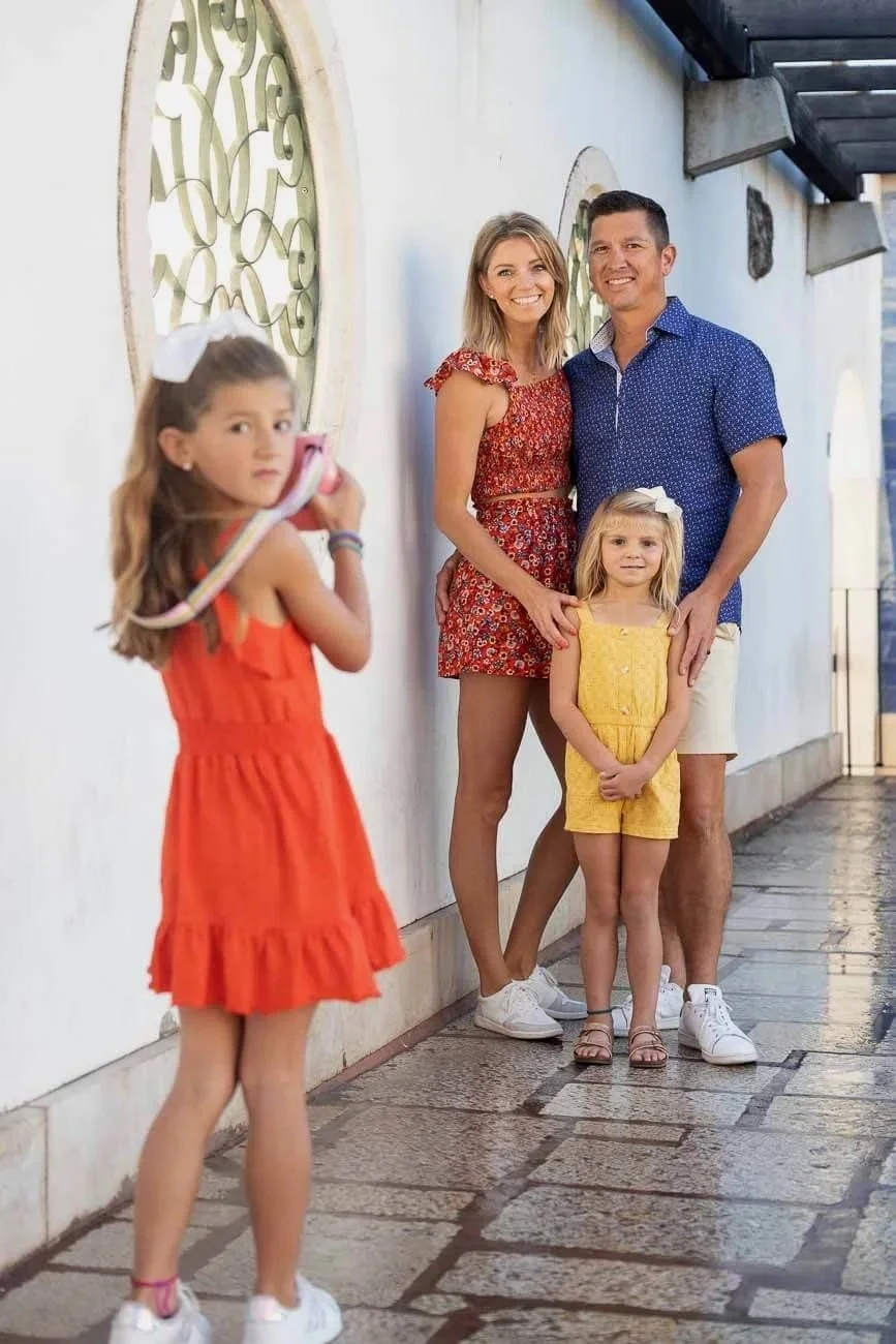 A family of four posing for a photographer in Lisbon. The mother and father stand close together, with the mother wearing a red floral dress and the father in a blue patterned shirt and shorts. Two young girls, one in a yellow dress and the other in 