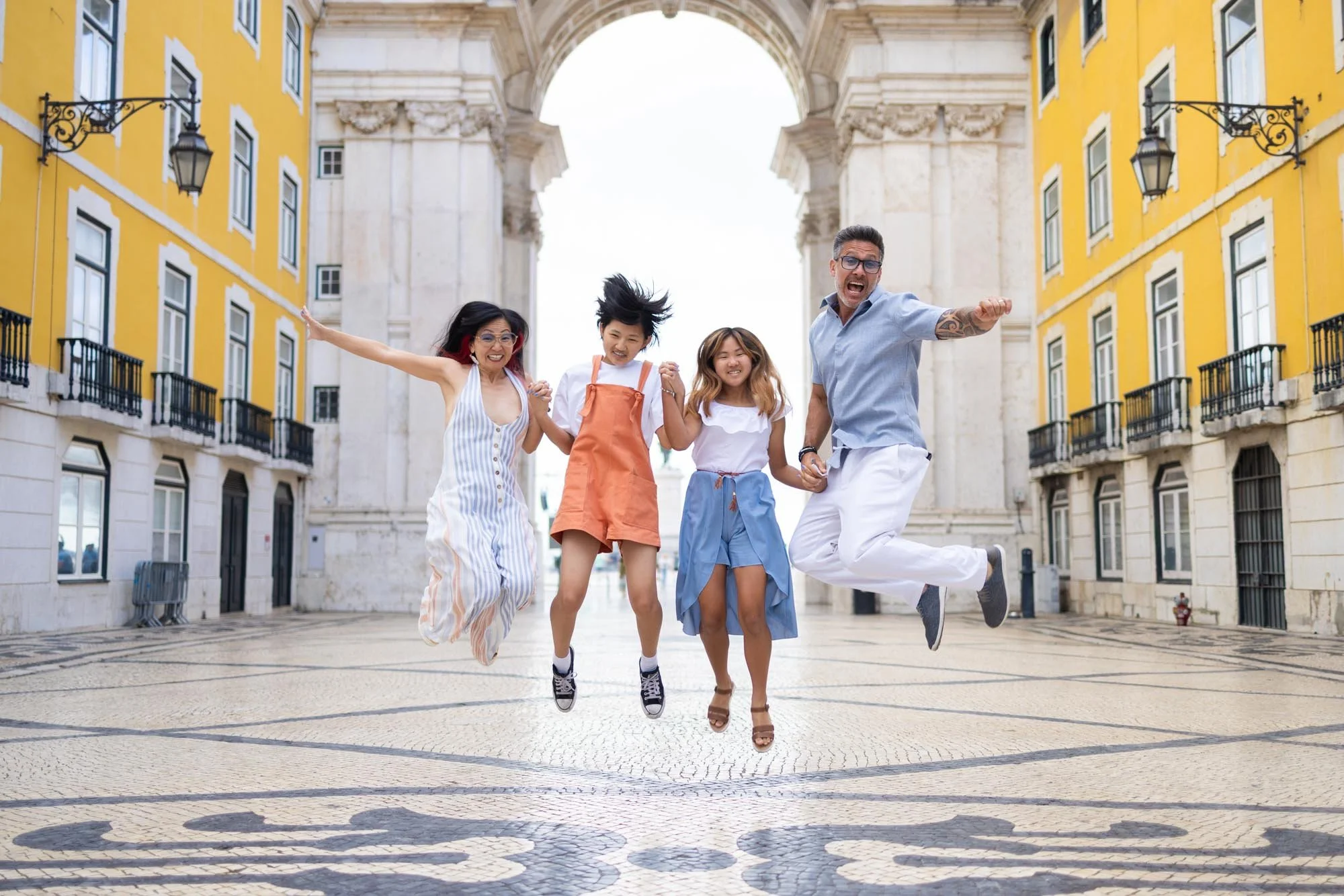 Four people of diverse backgrounds jumping and smiling in a European-style plaza with yellow buildings and an arch in the background.
