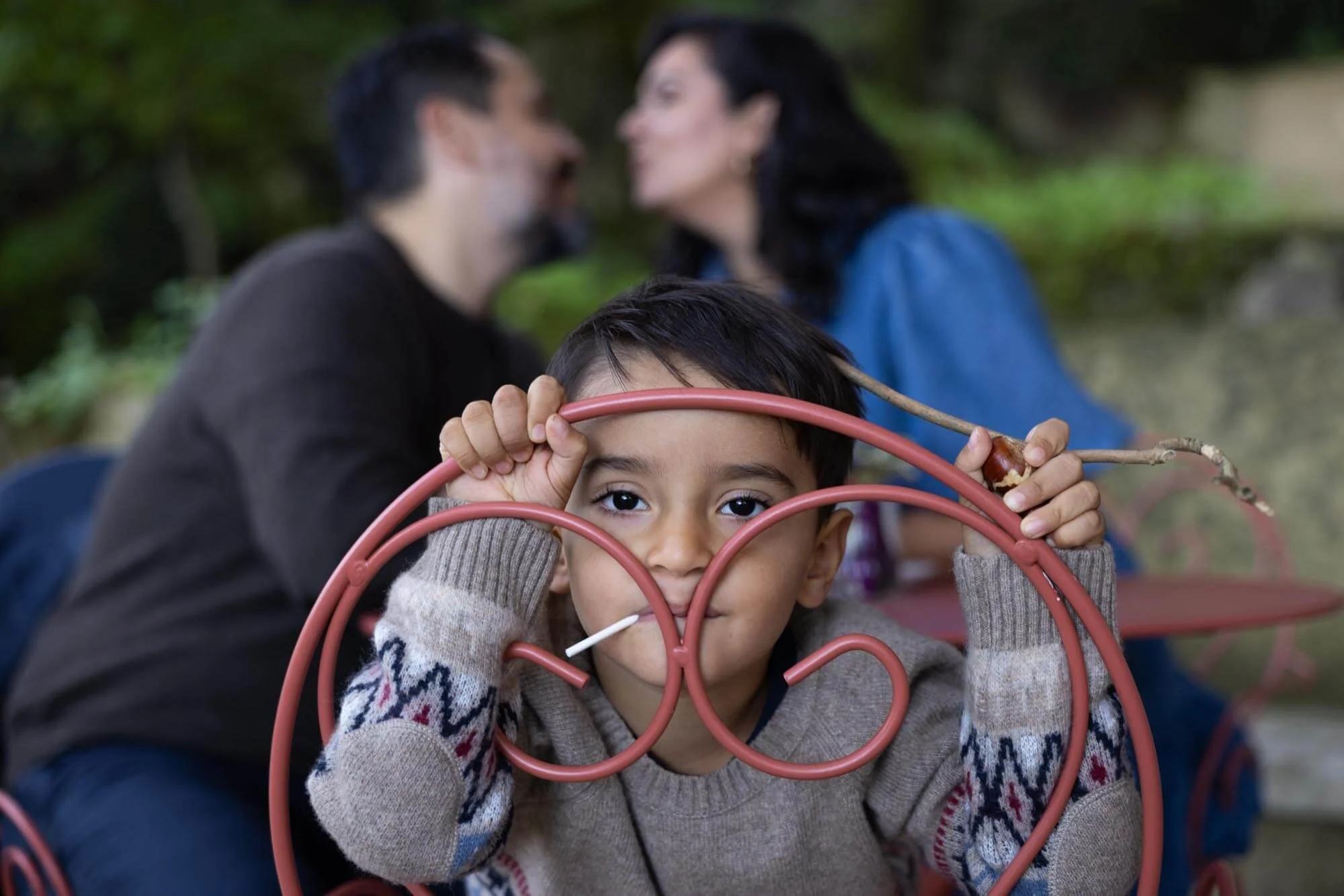 A young boy with dark hair and brown eyes looking through red metal chairs, with two adults blurred in the background, leaning in close to each other and smiling, outdoors in a natural setting.