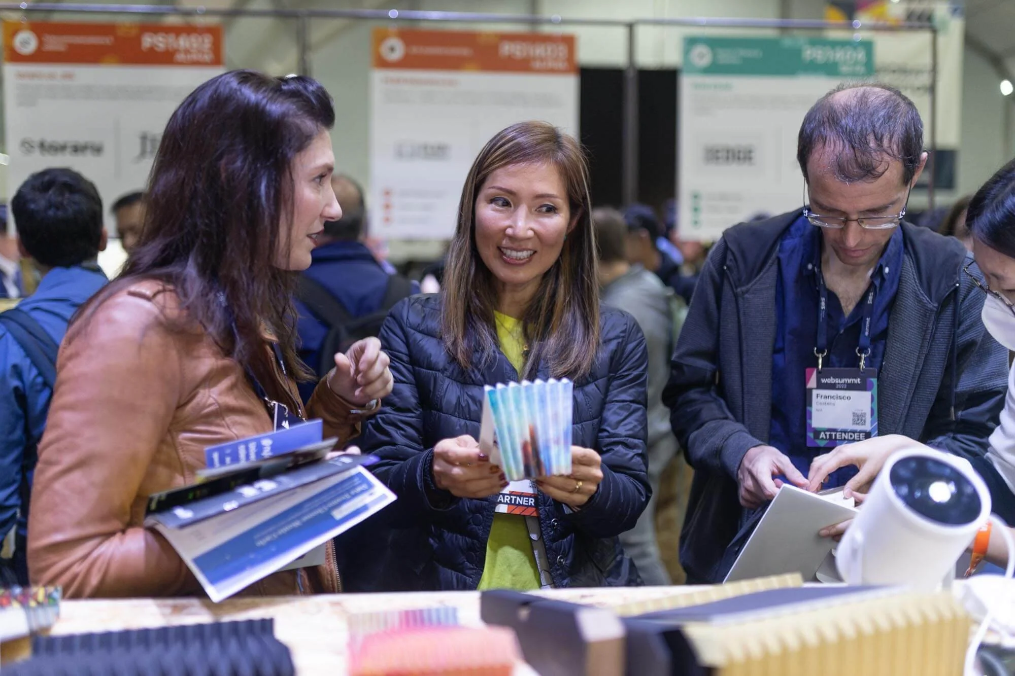 A group of people at an indoor conference or convention, engaging with displayed products and reading materials, with large informative posters in the background.