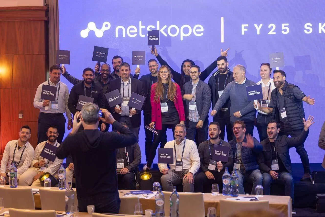 A group of people gathered on a stage at a conference, smiling and holding signs that say 'Own the Moment.' A person in front is taking a photo of them, with a large screen behind displaying the NetSkope logo and event information.