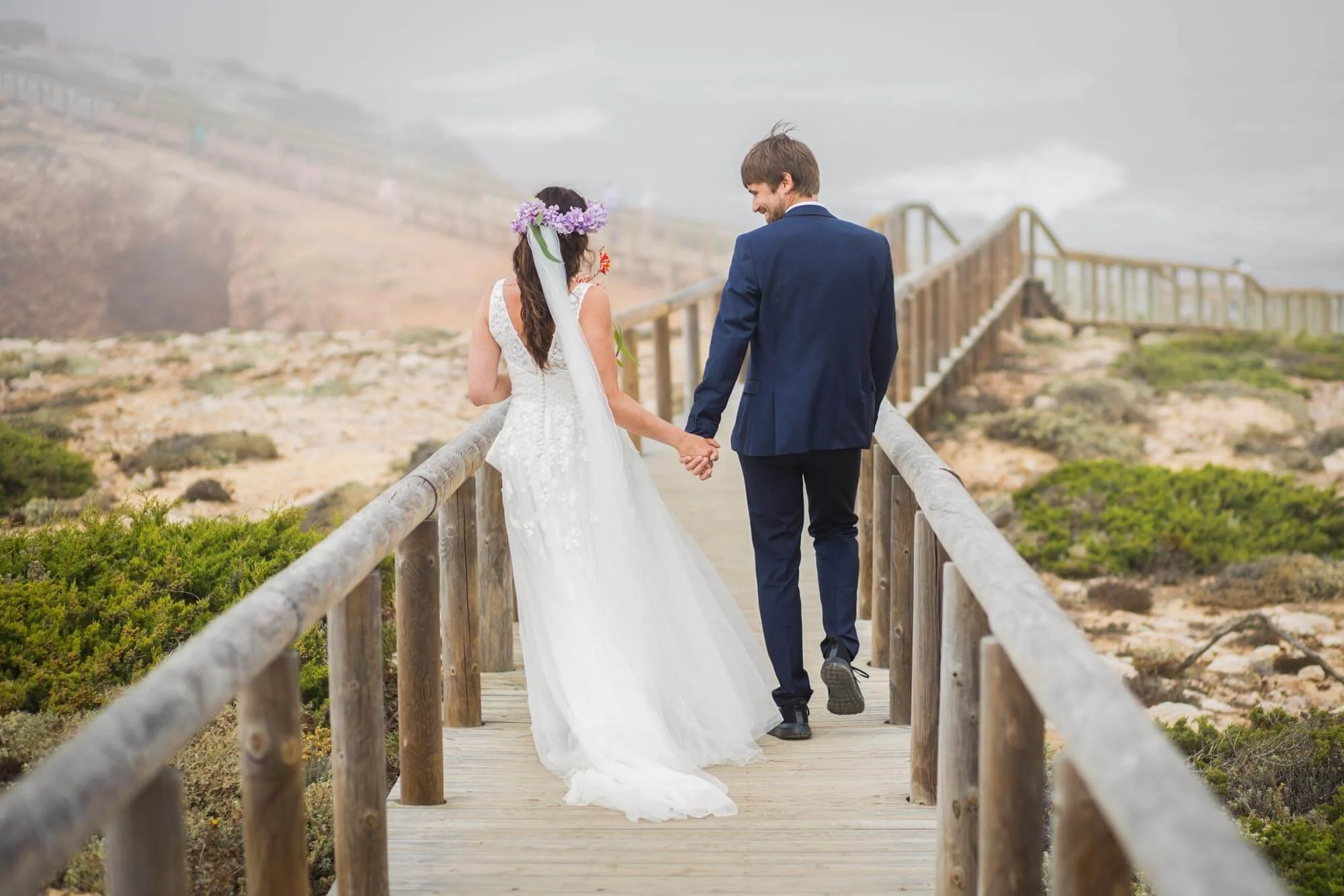 A bride and groom walking hand in hand on a wooden path outdoors, with a cloudy sky and greenery around.