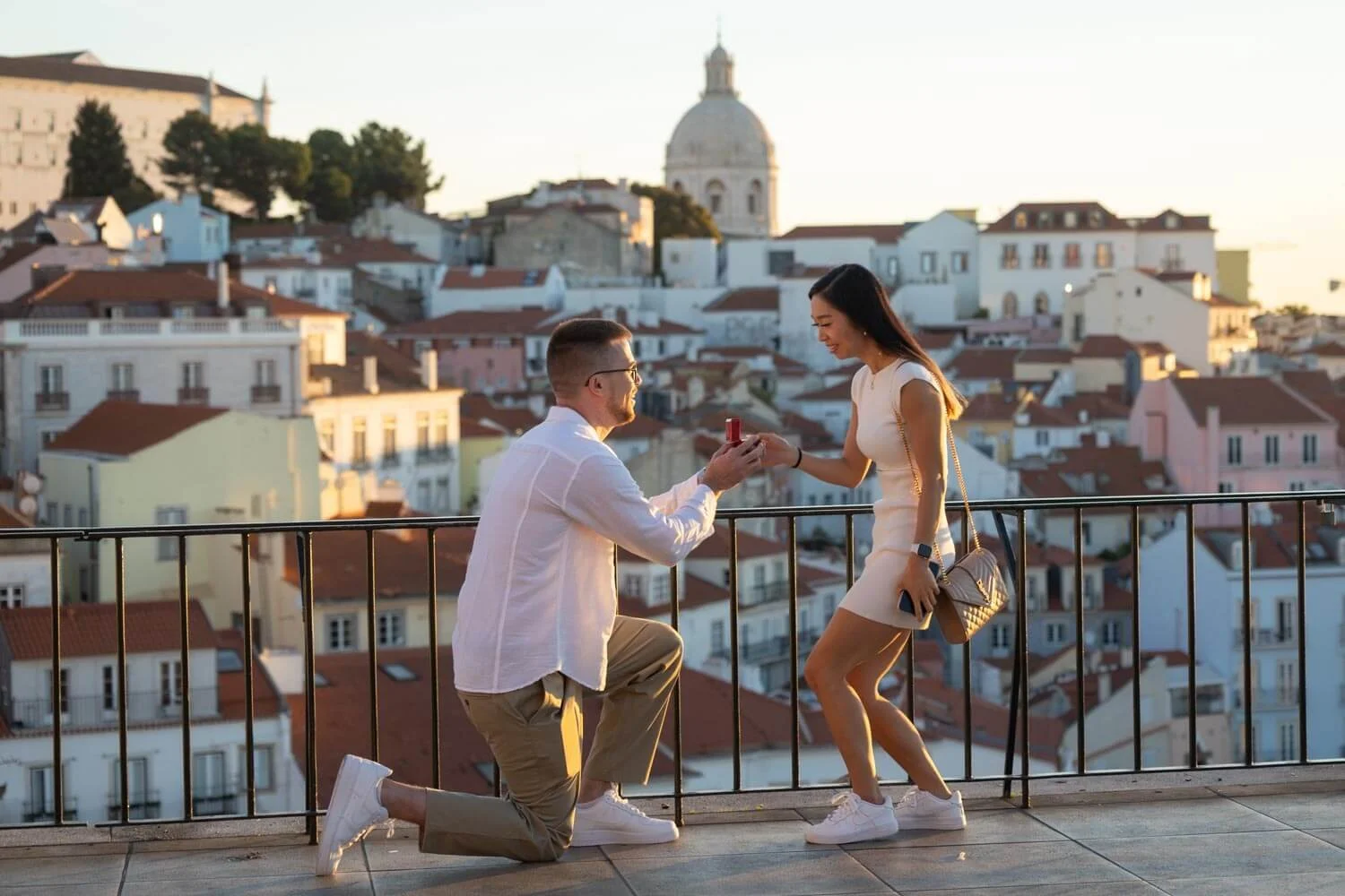 A man proposing marriage to a woman on a rooftop with a cityscape and historic buildings in the background during sunset.