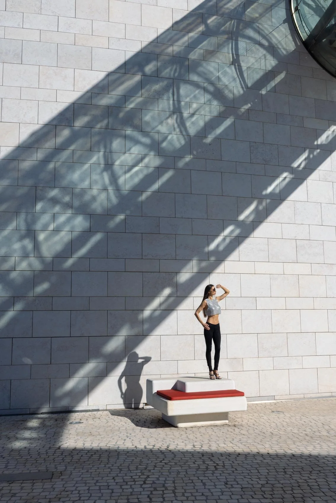 Woman posing on a modern white bench with a red cushion against a tiled wall and dramatic architectural shadows