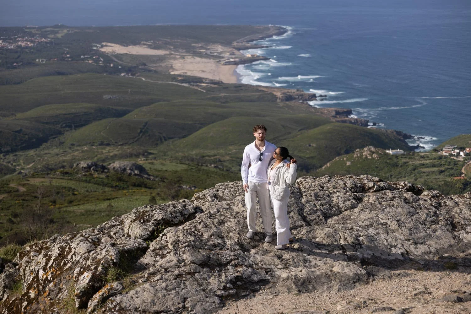 A couple dressed in white standing on a rocky hilltop overlooking a lush green coastal landscape with cliffs, beaches, and the ocean in the background.
