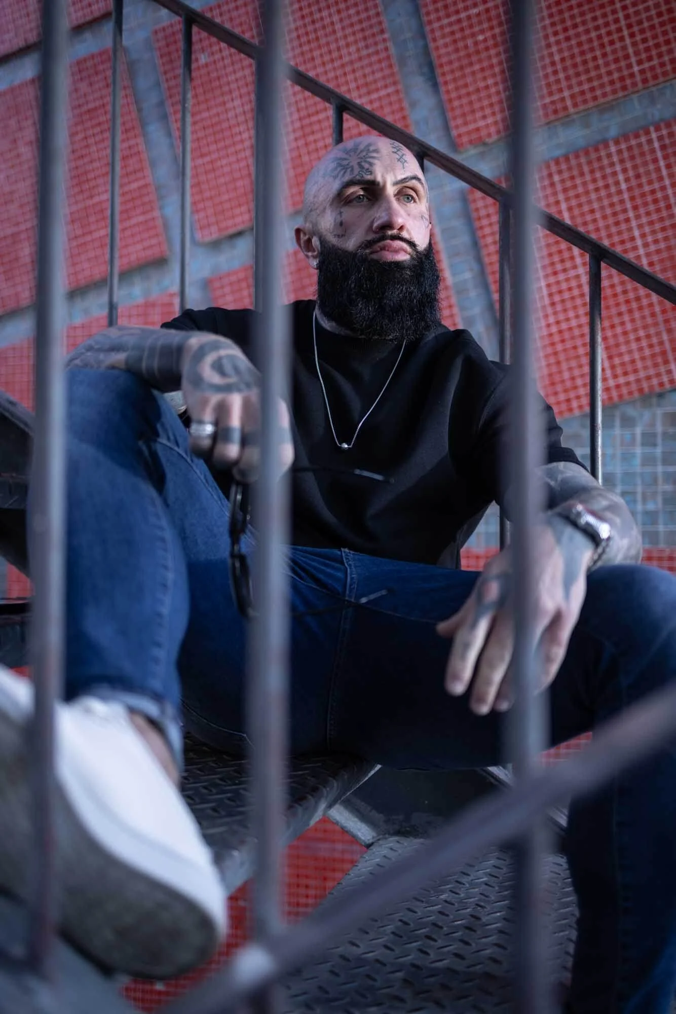 A seated man with a beard and tattoos, wearing a black shirt and jeans, is sitting on a metal staircase, looking off to the side. The background features a red and grey brick wall. The guy is posing for a photographer in Lisbon downtown.