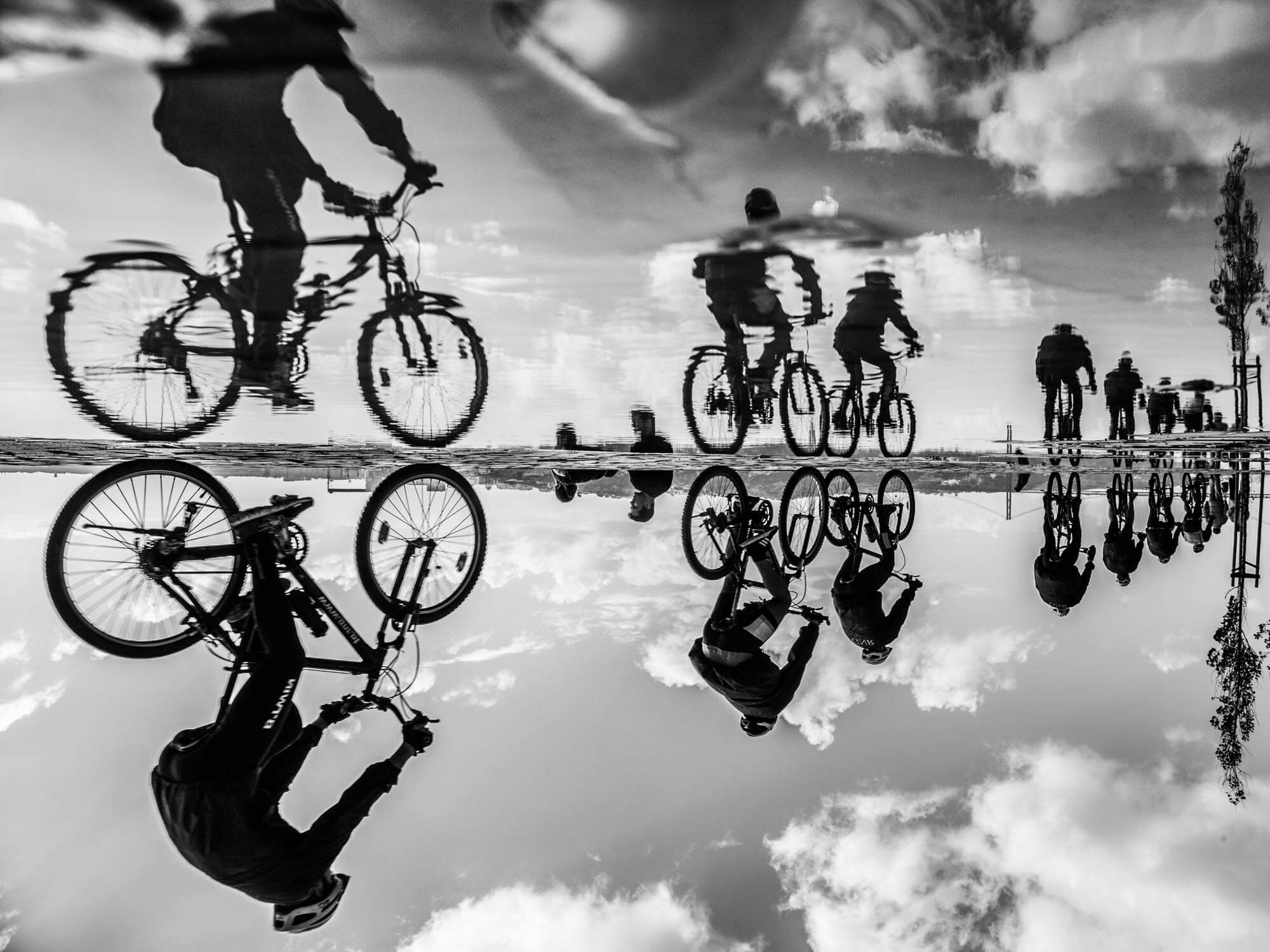 Reflection of people riding bicycles and walking, with some wearing helmets, in a large puddle or shallow water, creating a mirror image with clouds and sky above.