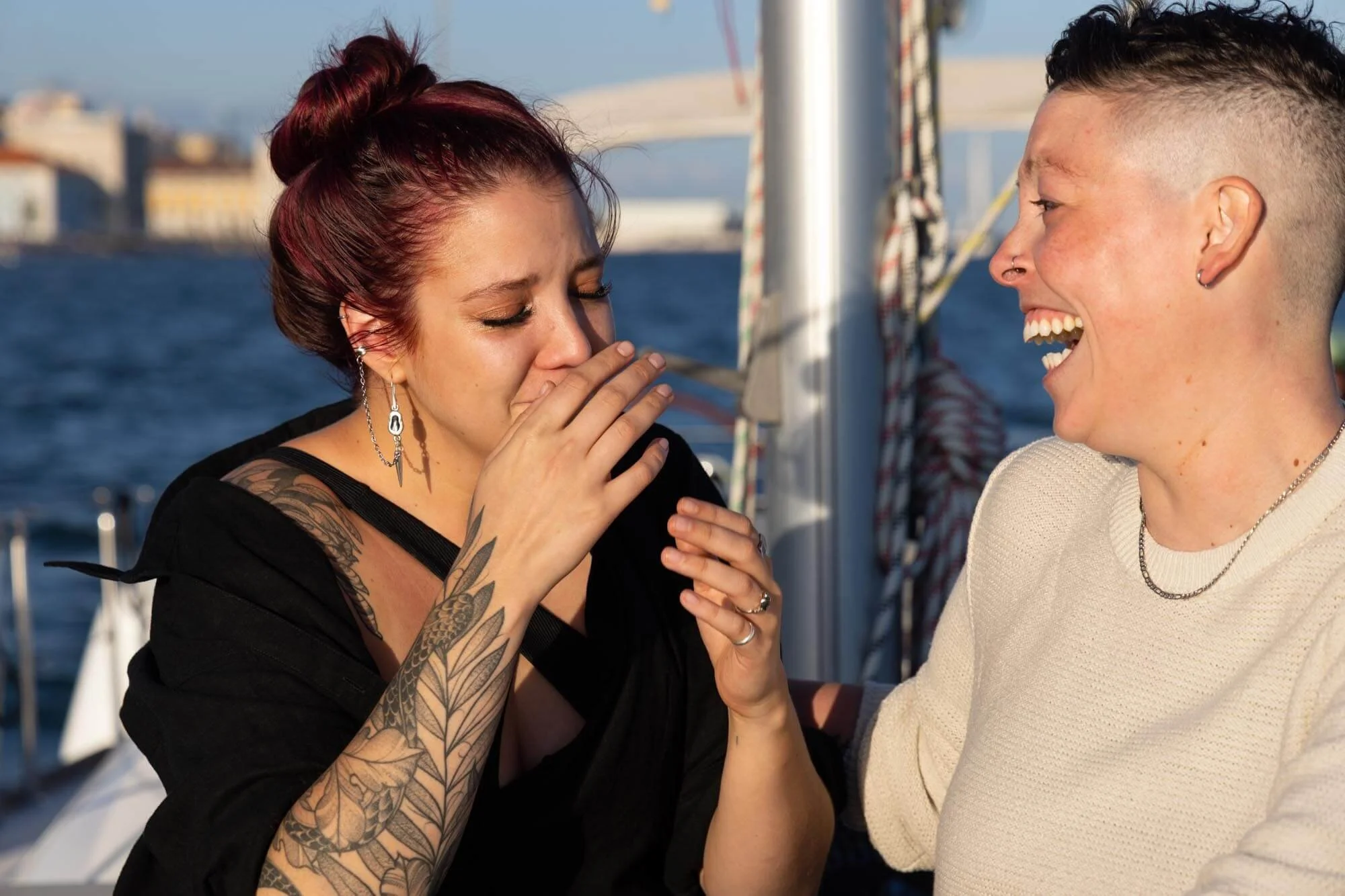 Two women on a boat share an emotional moment, one woman is crying and the other woman is laughing, with water and buildings in the background.