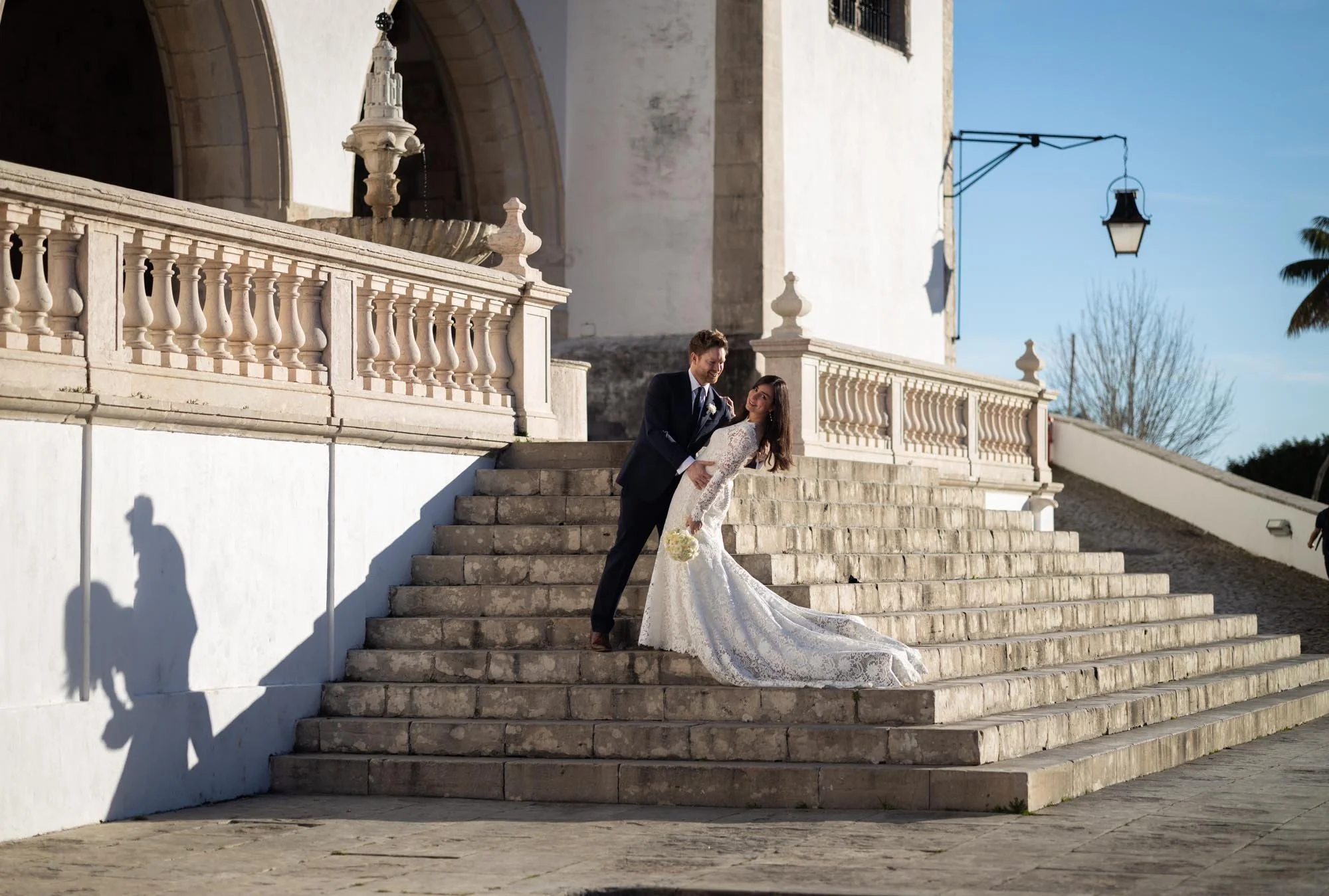 A bride and groom are dancing on stone stairs outside a white building, with a large fountain and a street lamp visible in the background, during a sunny day.
