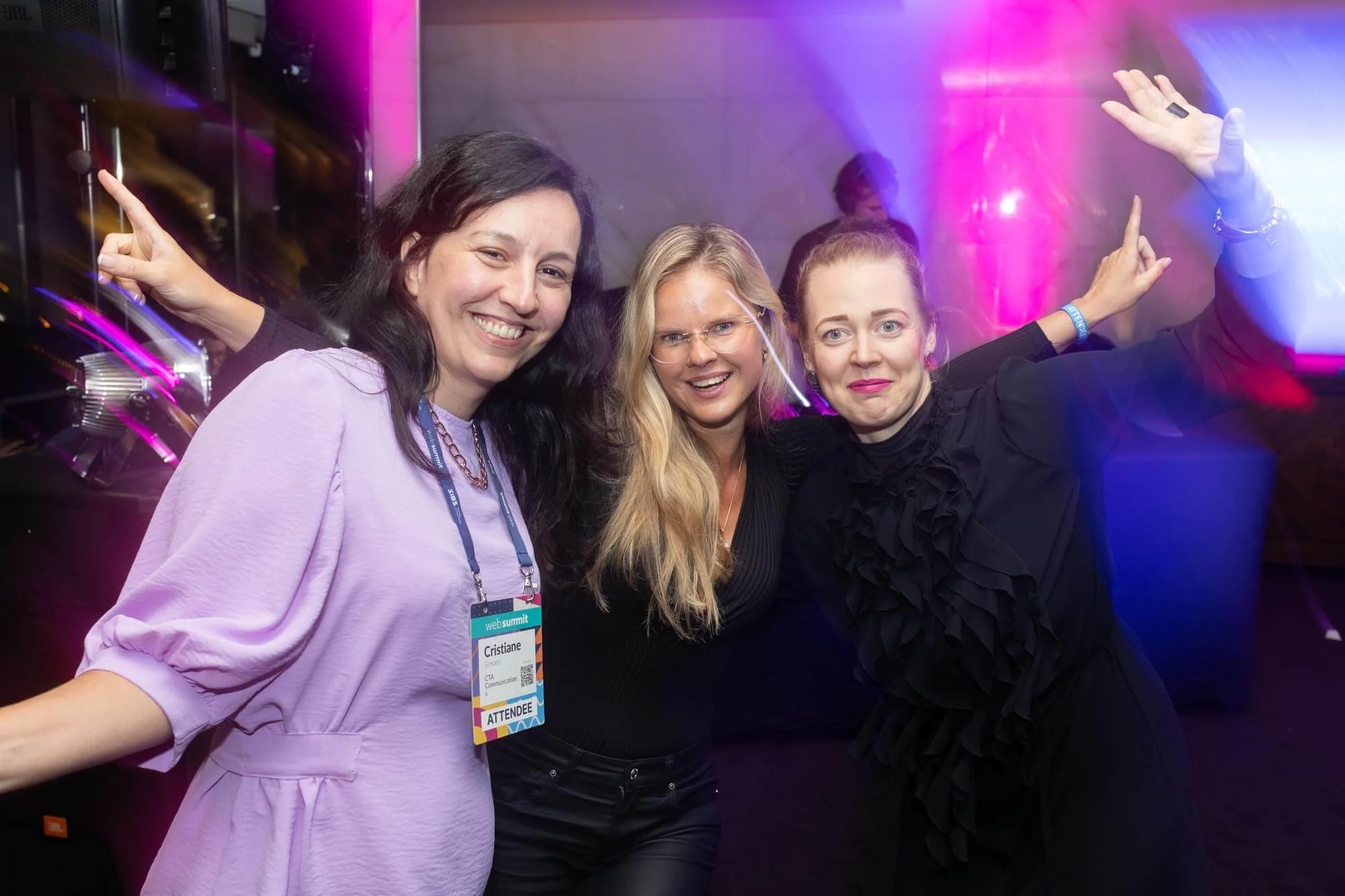 Three women smiling and dancing at an event, with colorful purple and blue lighting in the background.