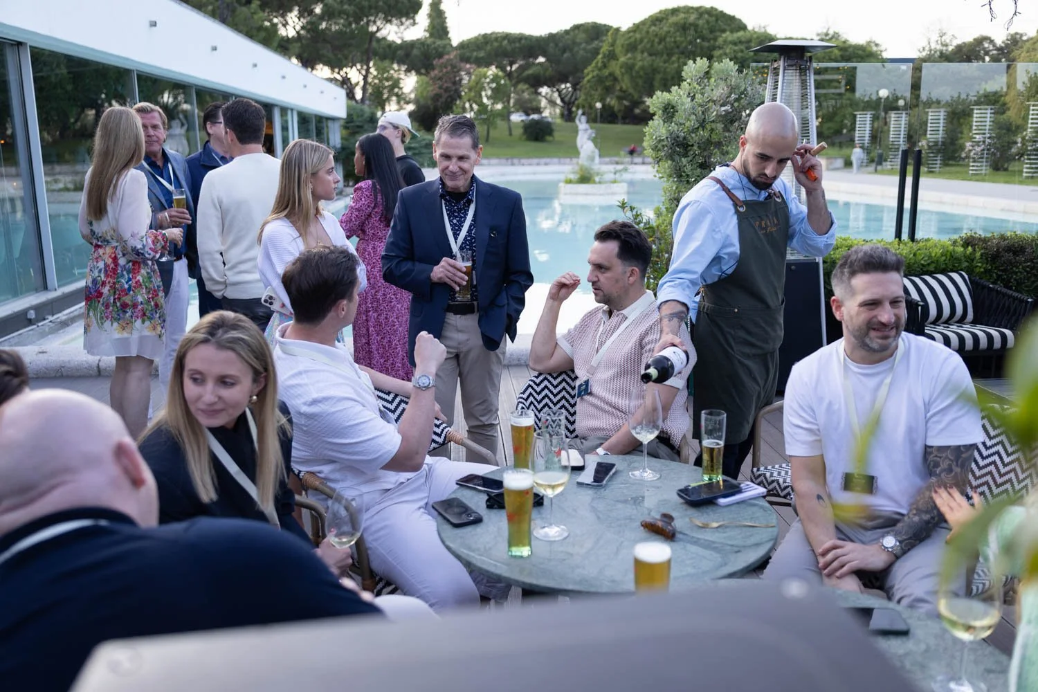 People socializing at an outdoor corporate dinner event in Lisbon,  near a pool, with drinks in hand, some seated and some standing, and a waiter serving drinks.