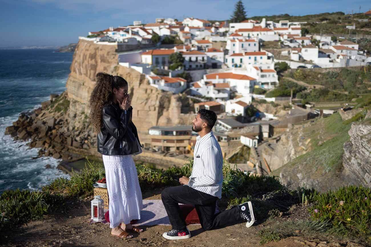 A man kneeling on one knee proposing to a woman on a scenic overlook with cliffs and a coastal town in the background.