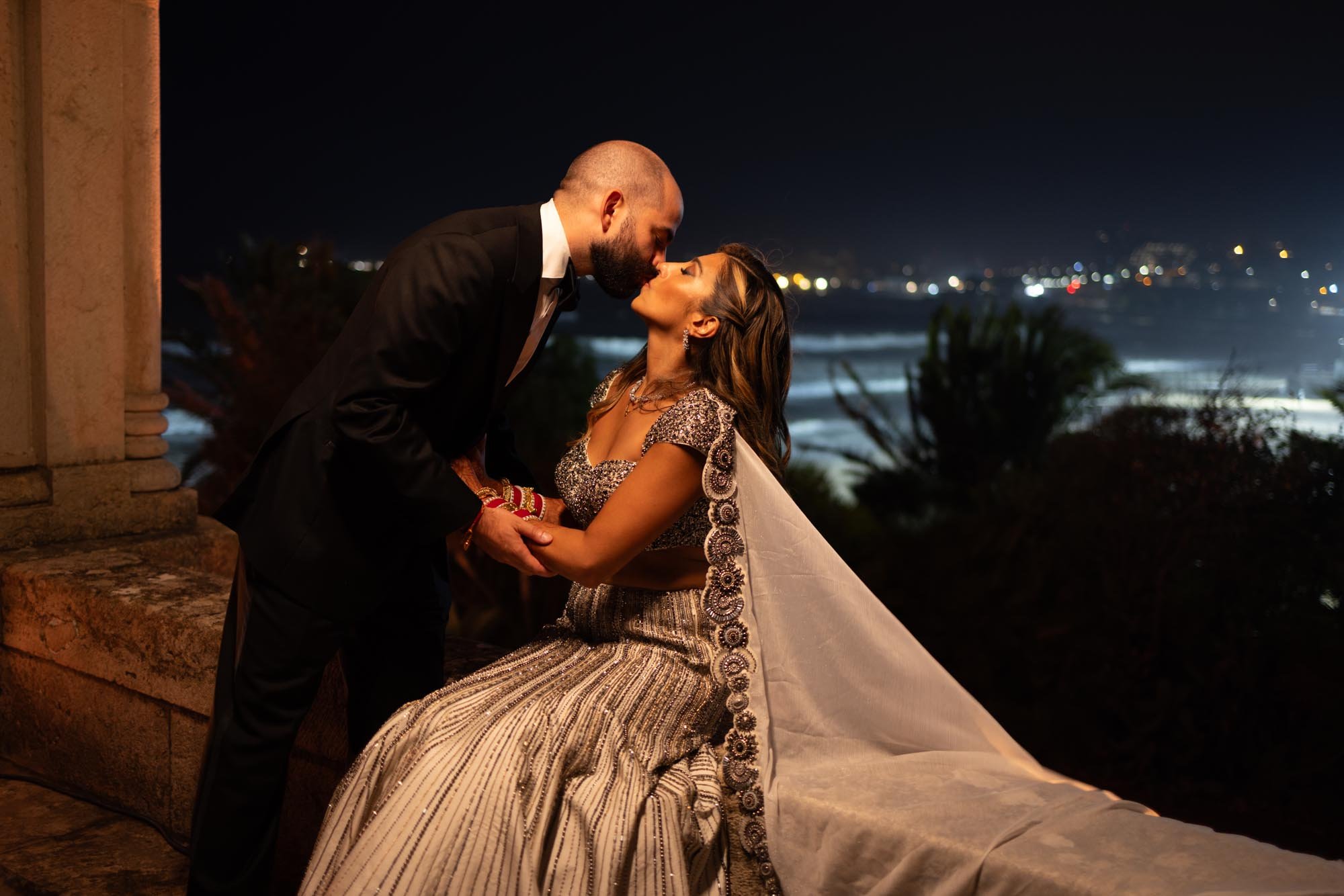 A couple dressed in formal attire sharing a romantic kiss at night with a cityscape and ocean in the background.