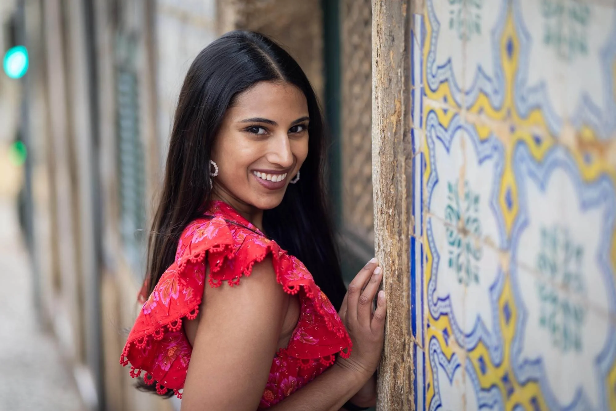 A woman with long dark hair, smiling, wearing a red, ruffled dress, standing beside a mural wall with colorful patterns.