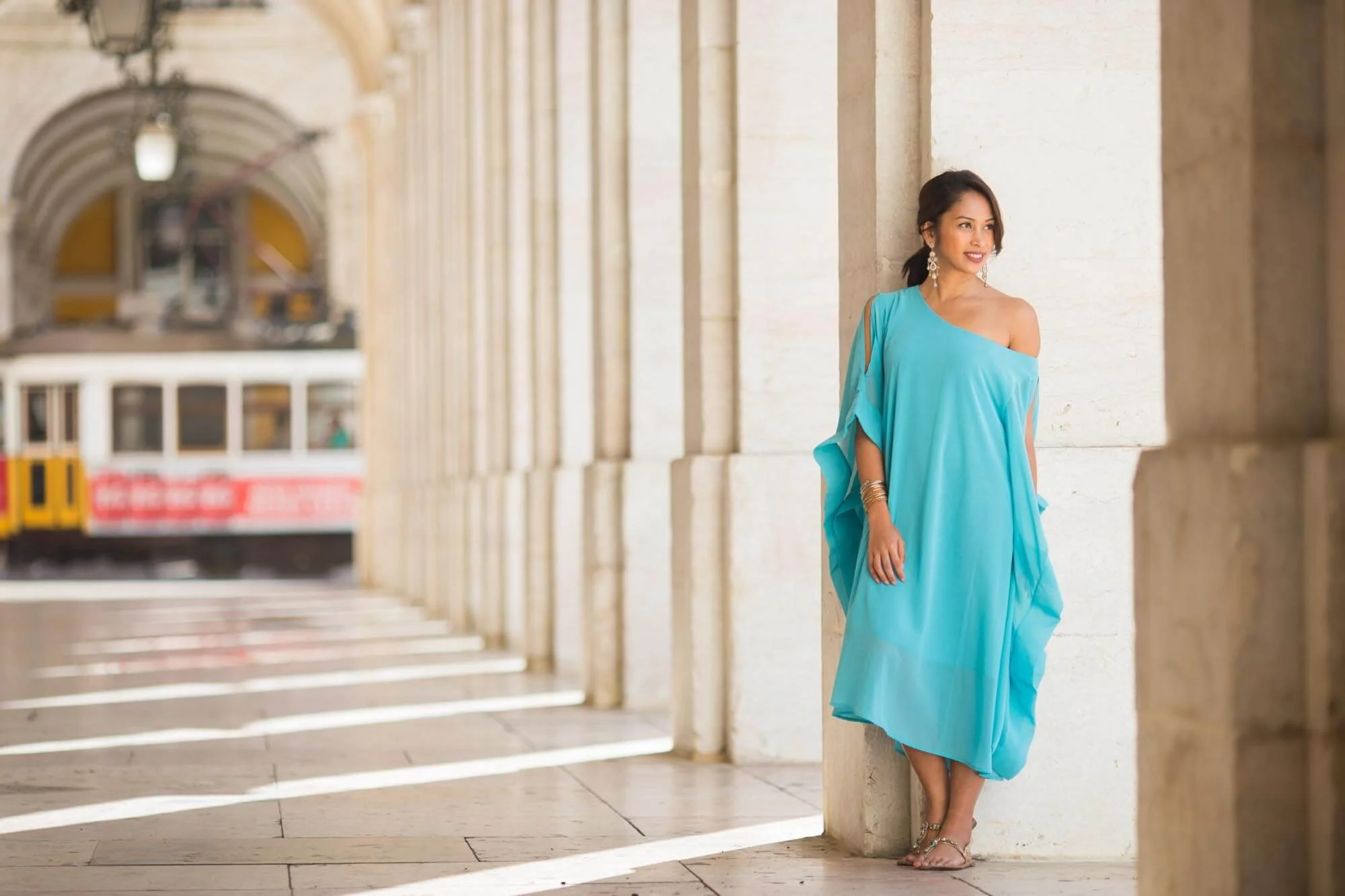 A woman in a turquoise dress with off-the-shoulder detail standing in a corridor with stone columns, with a tram in the background.