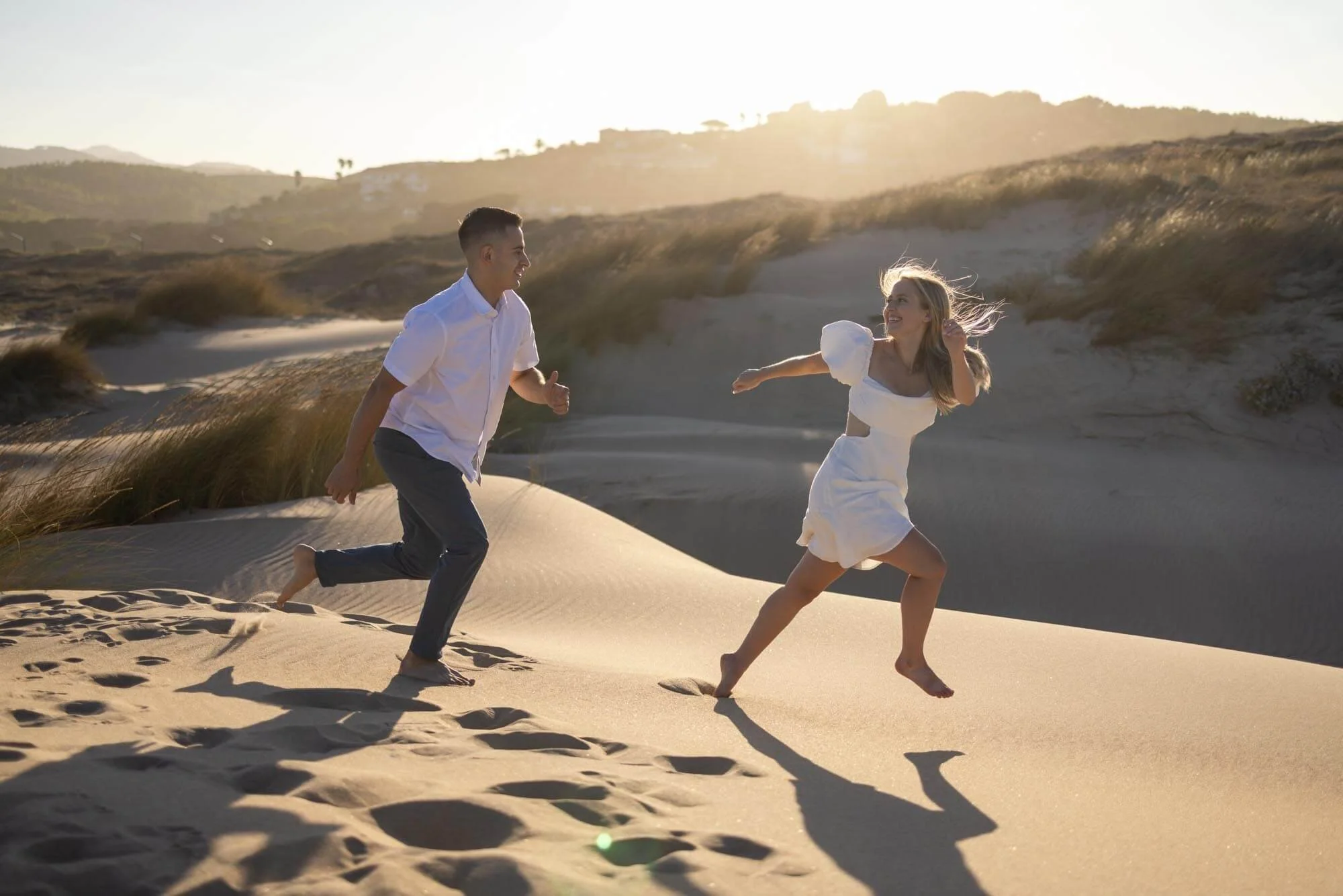 A young man and woman running barefoot on sand dunes, laughing, with the sun setting behind them.