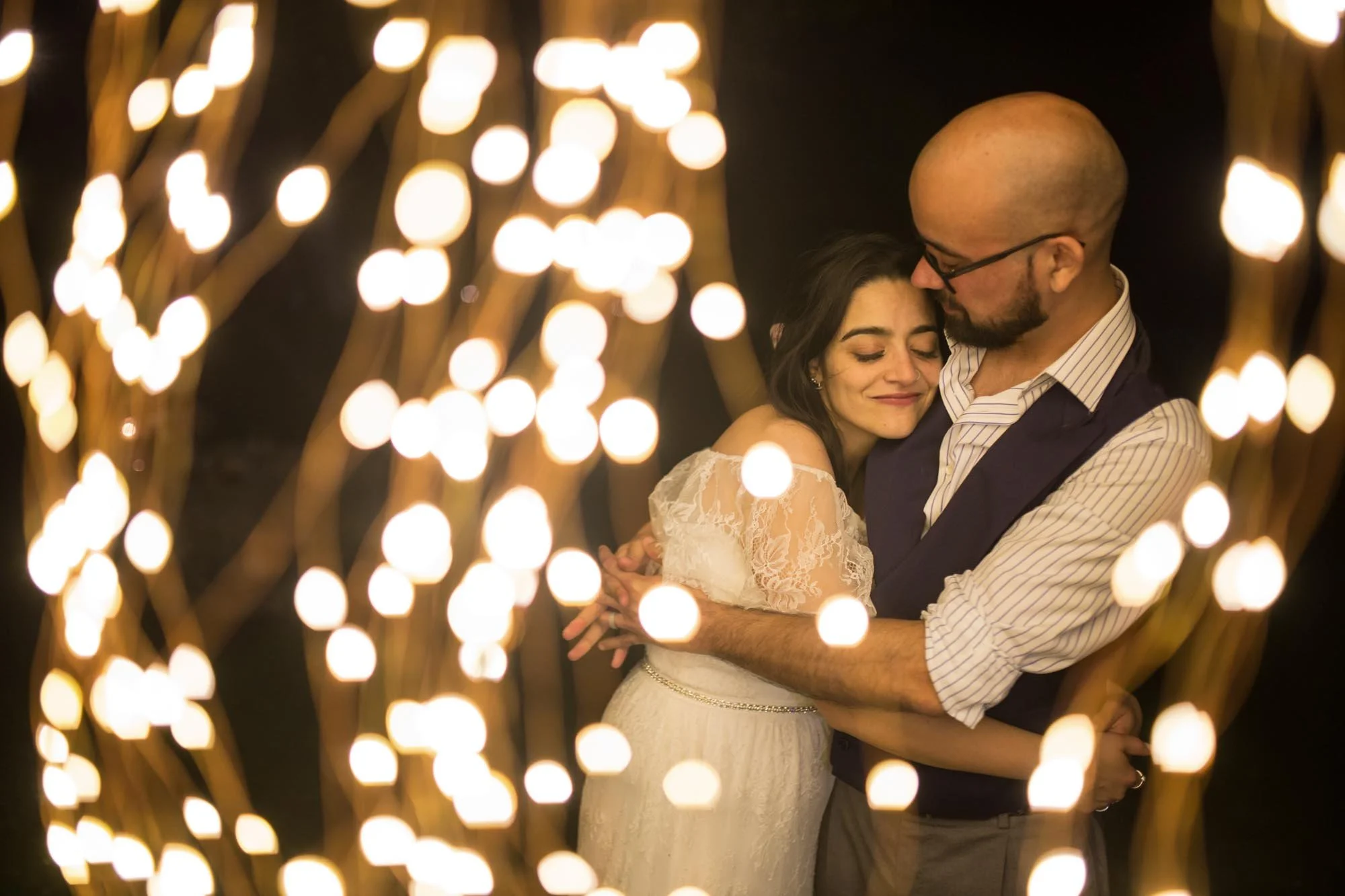A couple embracing at night, with blurred string lights in the foreground, celebrating a special occasion.