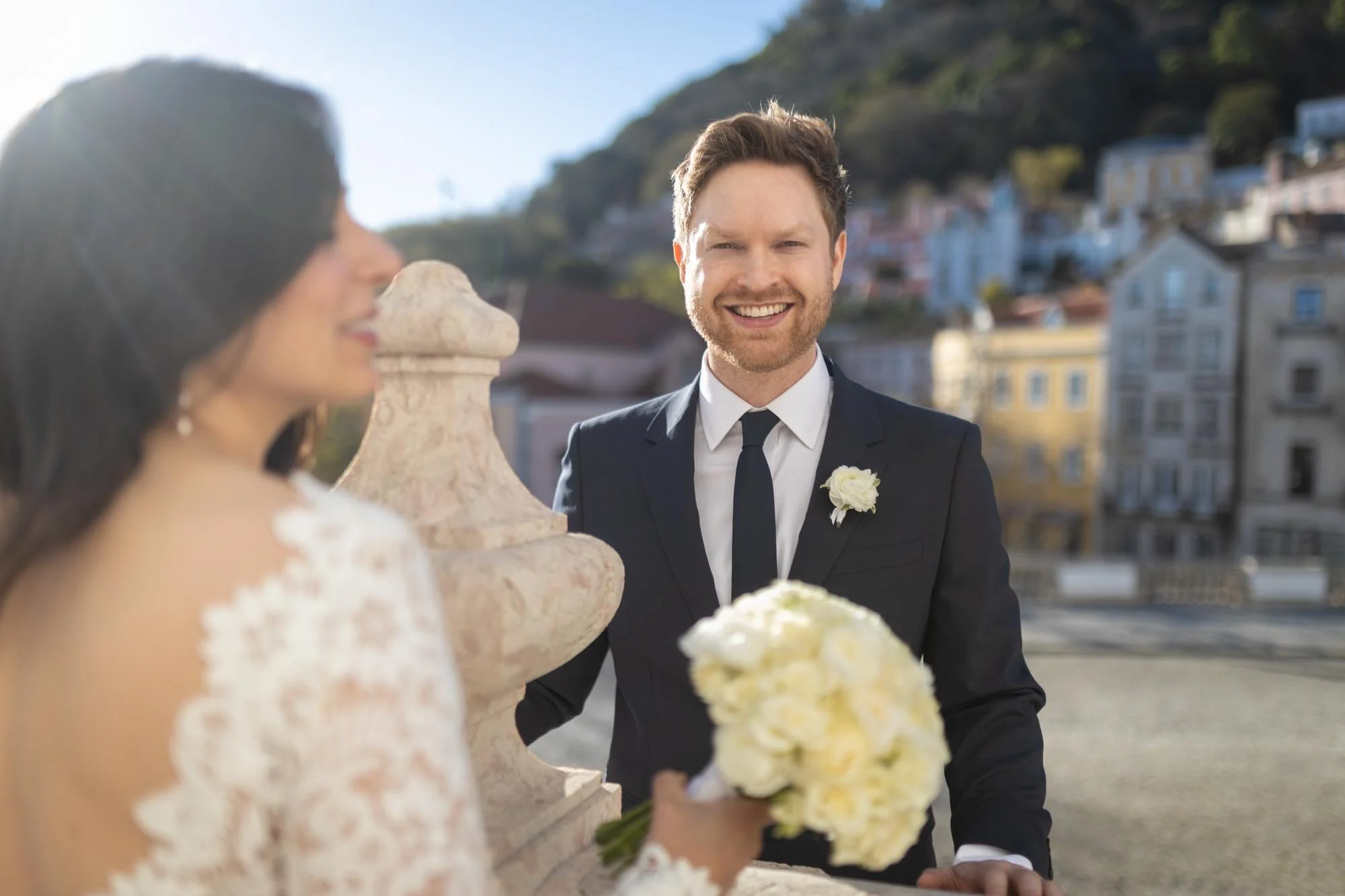 A smiling man in a black suit, white shirt, and black tie, standing outdoors on a sunny day with a cityscape in the background. He has a white flower boutonniere on his suit. A woman in a lace dress holds a bouquet of white flowers, with her face blu