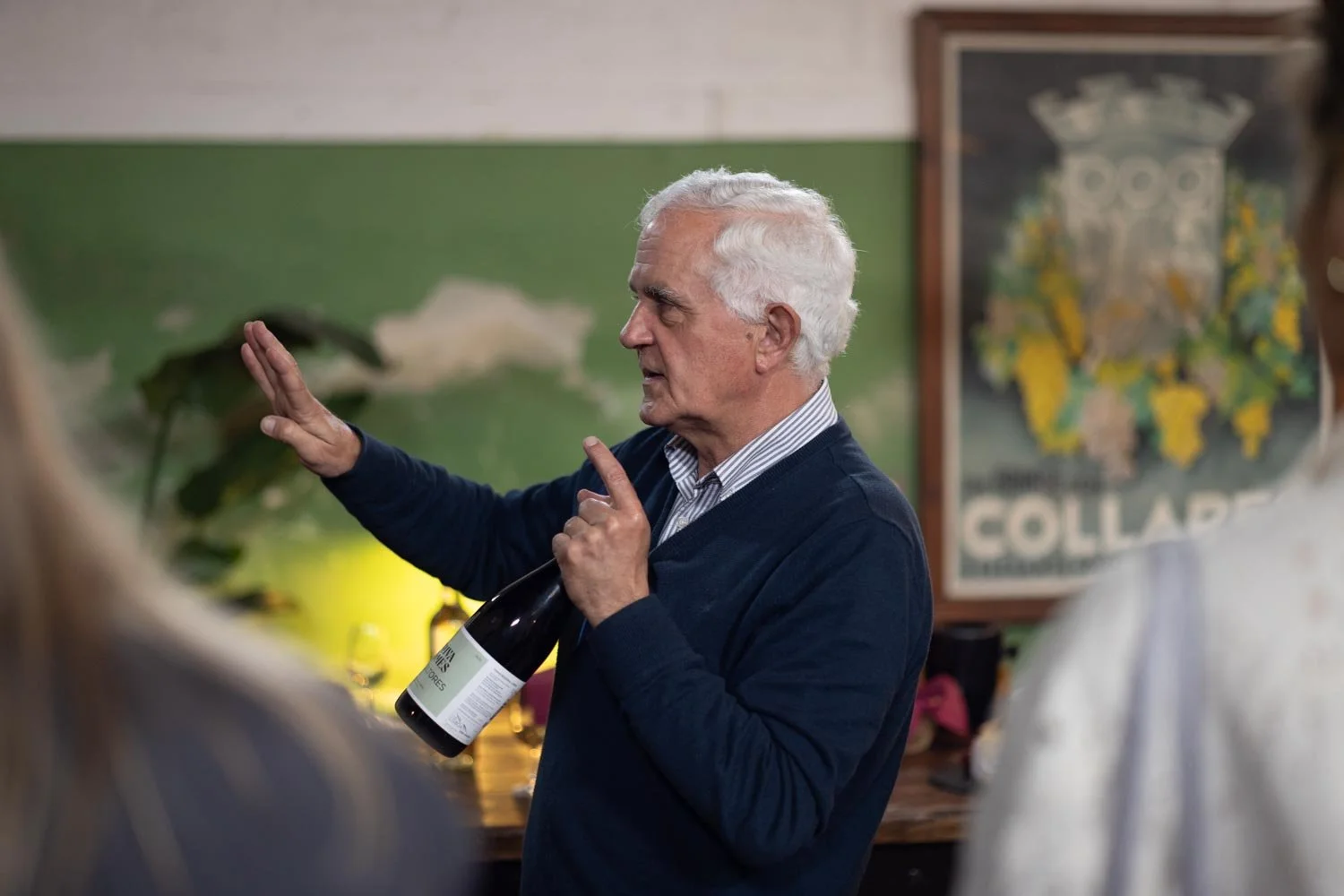 Wine tasting activity for a corporate retreat in Lisbon. An elderly man with white hair gesturing with his hand while holding a wine bottle, speaking to a group of people in a room with green and white walls, with artwork and a plant in the backgroun