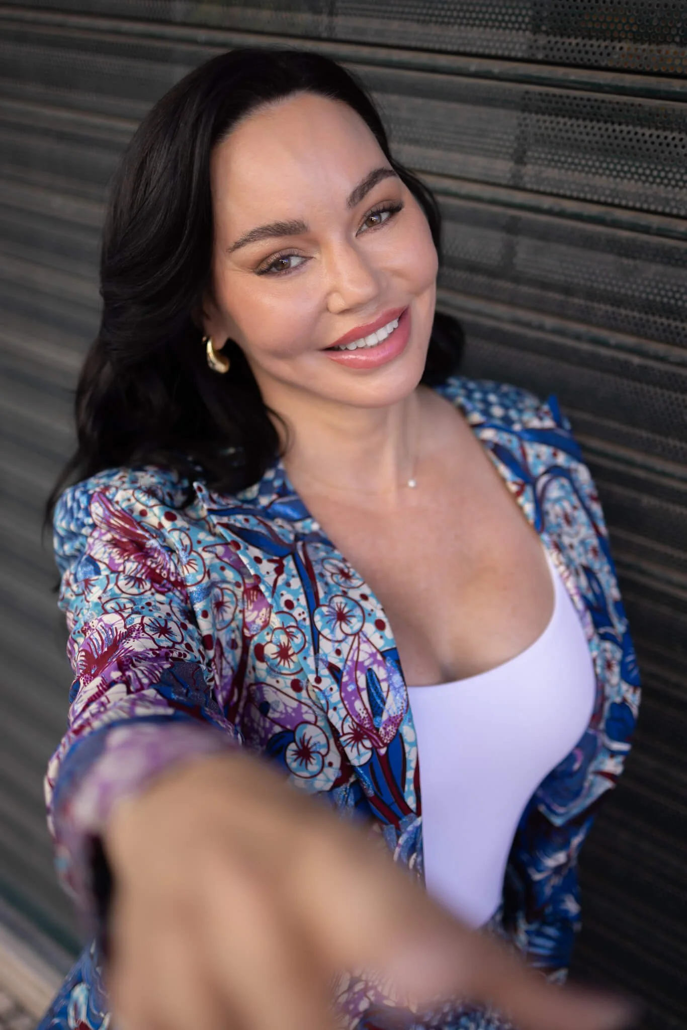 A woman with dark hair, wearing a patterned blue and purple jacket over a white top, taking a selfie with a smile.