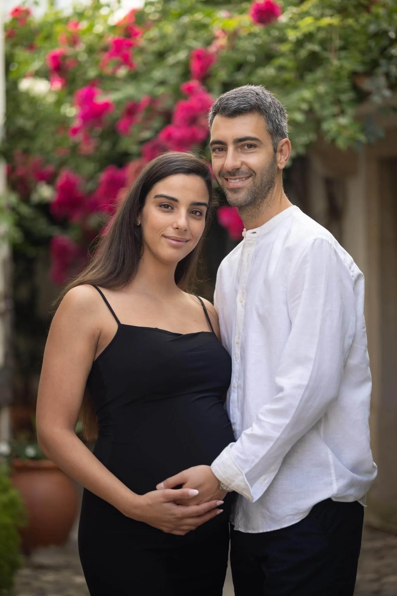 A pregnant woman with long dark hair wearing a black spaghetti strap dress, standing next to a man with short gray hair wearing a white button-up shirt, both smiling and holding hands, outdoors with pink flowers in the background.