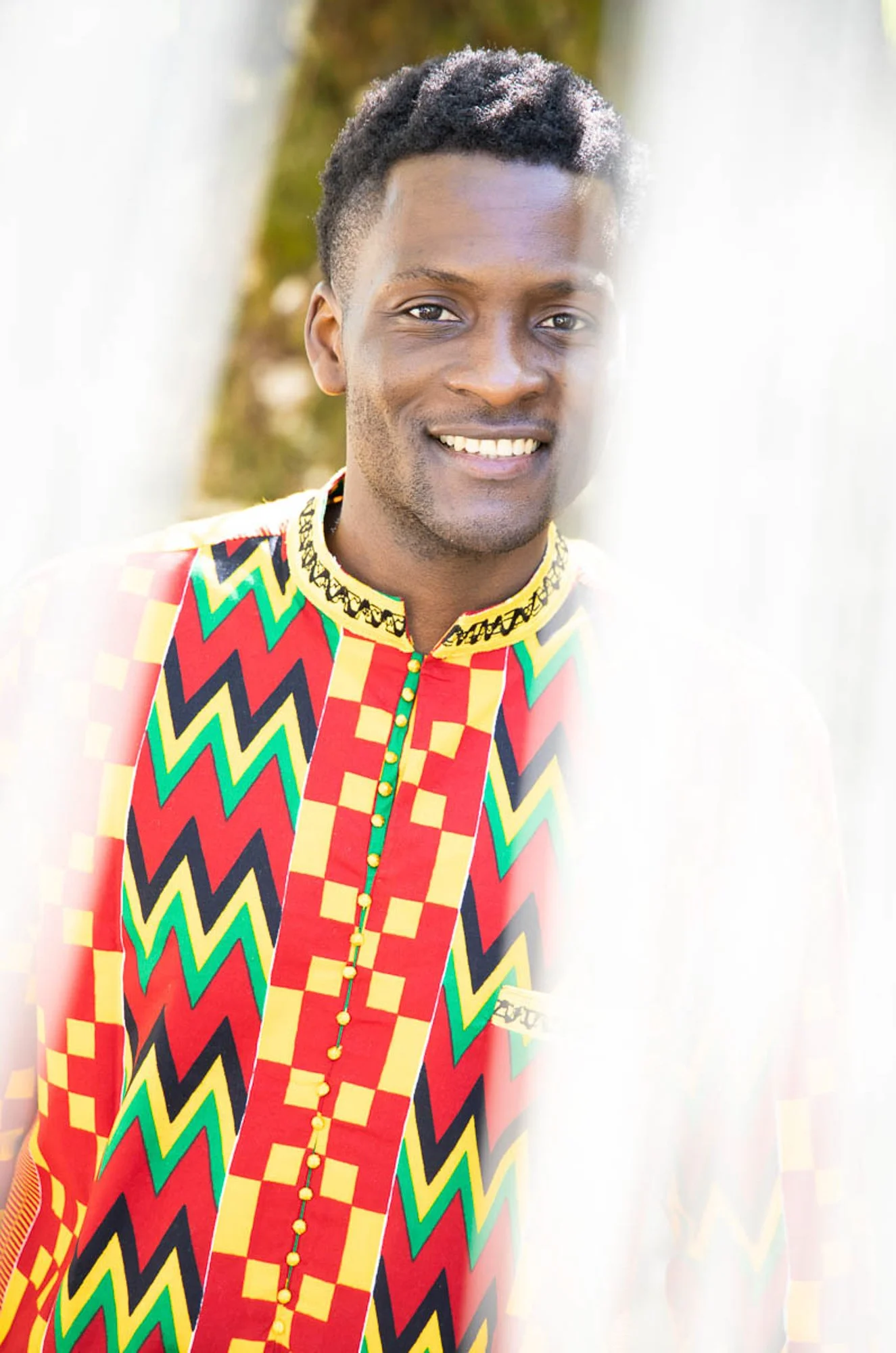 A smiling man with dark hair in a colorful African print shirt outdoors, with a bright background and soft focus.