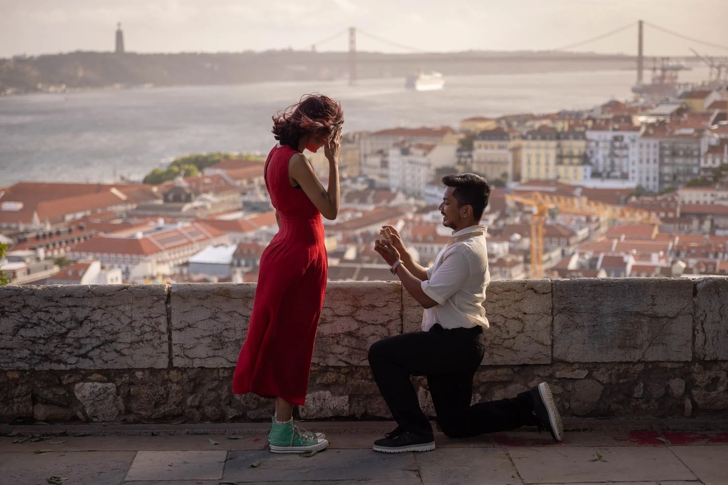 Sunset Secret Proposal Photoshoot at São Jorge Castle with a lovely couple from India. The guy is on the knees and proposing a girl in a red dress with a beautiful Lisbon city backdrop.
